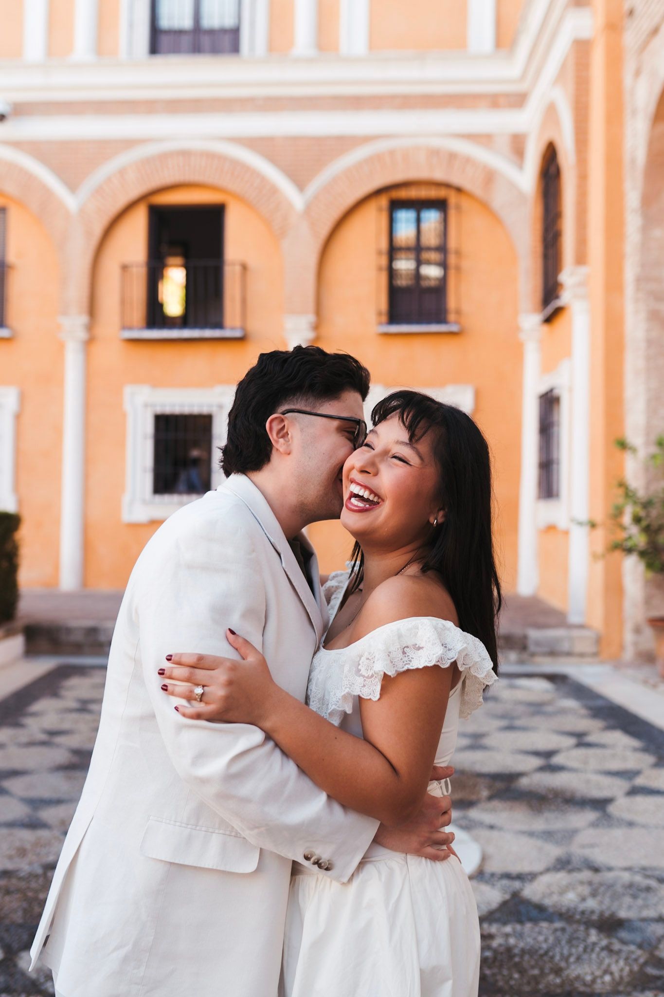 Asian couple laughing and embracing in front of golden arched building in Seville, Spain at dusk.