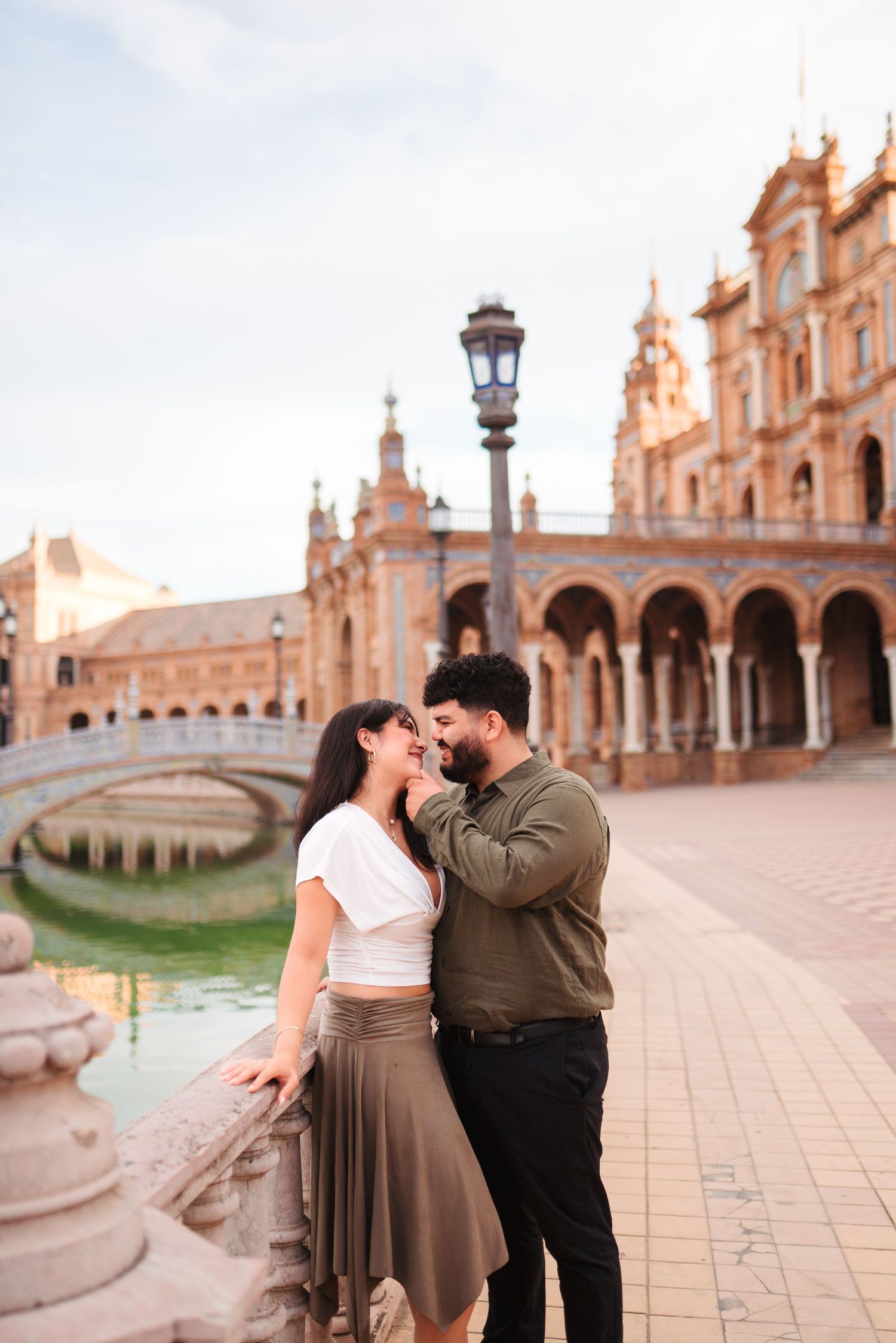 Romantic couple embracing at Plaza de España in Seville, Spain, with ornate architecture and canal in background.