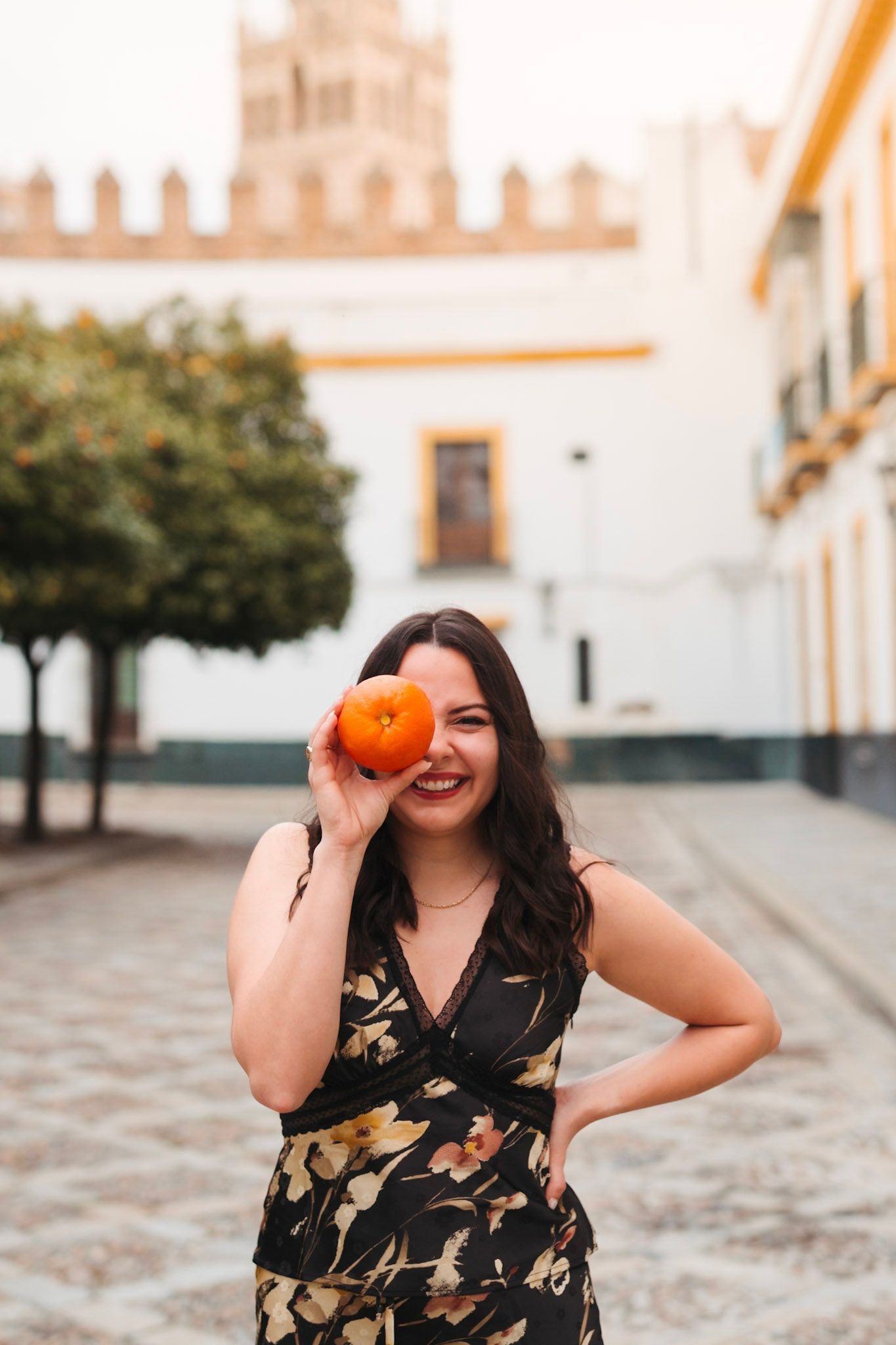 Smiling woman in floral dress holds orange over eye in sunny plaza.