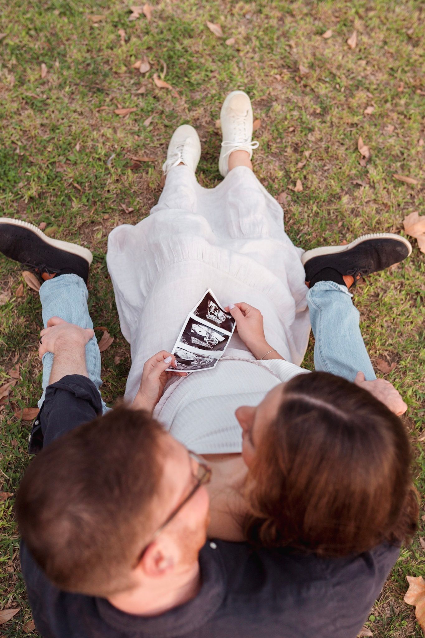 Couple sitting on grass holding ultrasound photos of their expected baby.
