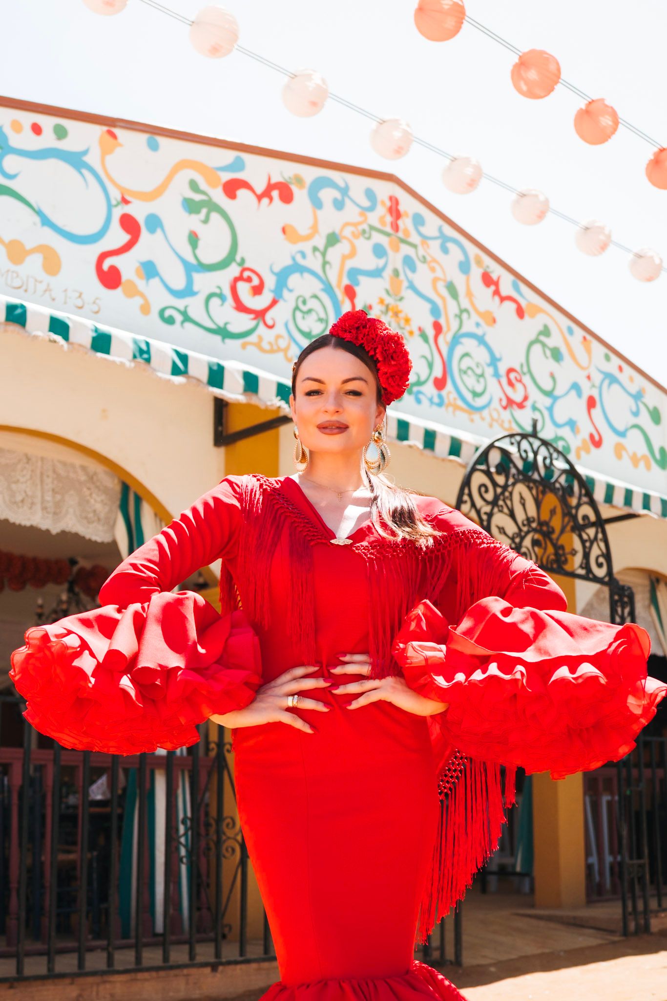 Flamenco dancer in red ruffled dress posing at colorful Seville fairground.