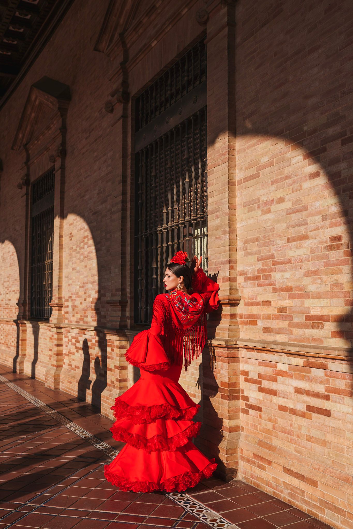 Flamenco dancer in red ruffled dress posing against brick wall with arched windows in Seville, Spain.