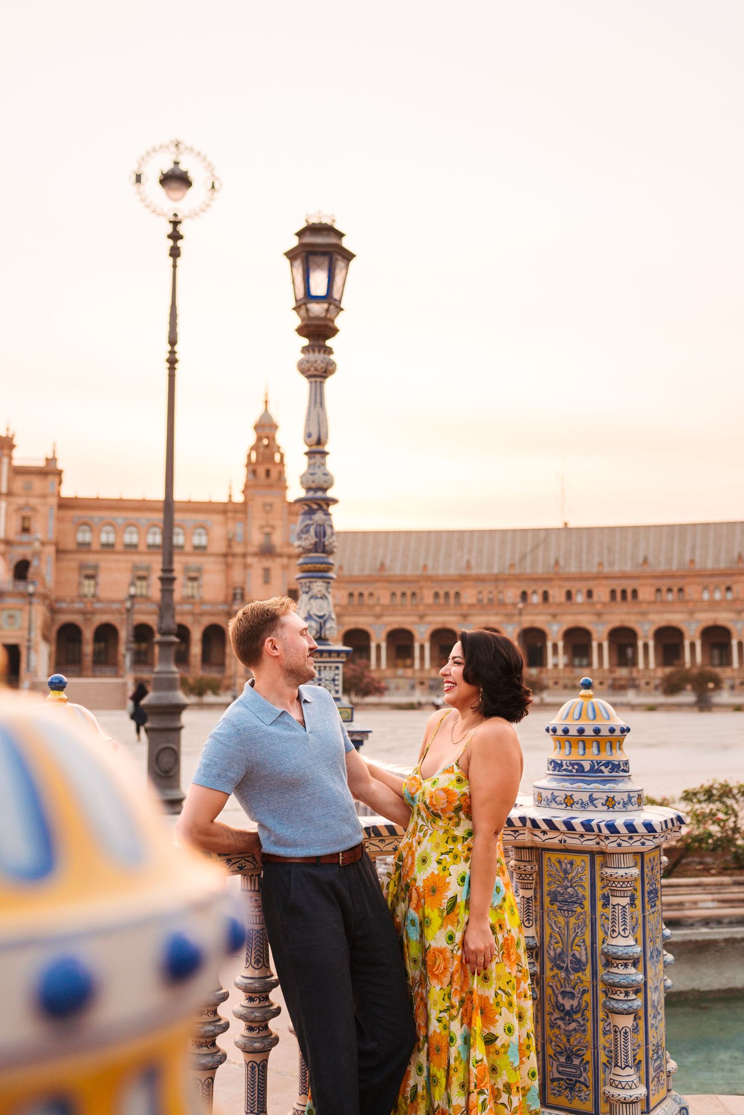 Couple smiling at each other at Plaza de España in Seville, Spain, surrounded by colorful tiles and a ornate lamppost.