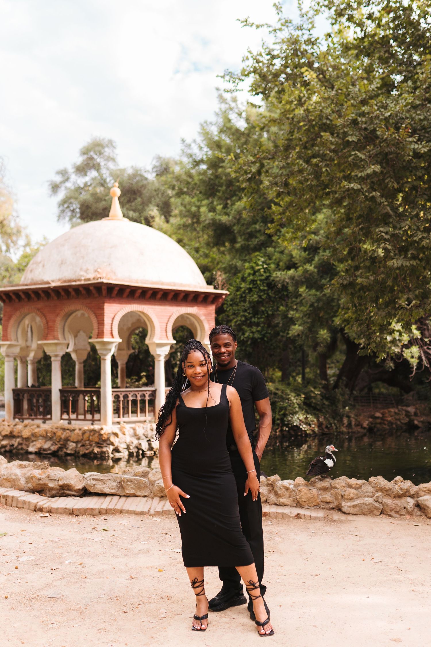 A couple in black outfits poses near an ornate pavilion surrounded by lush trees in a scenic park.