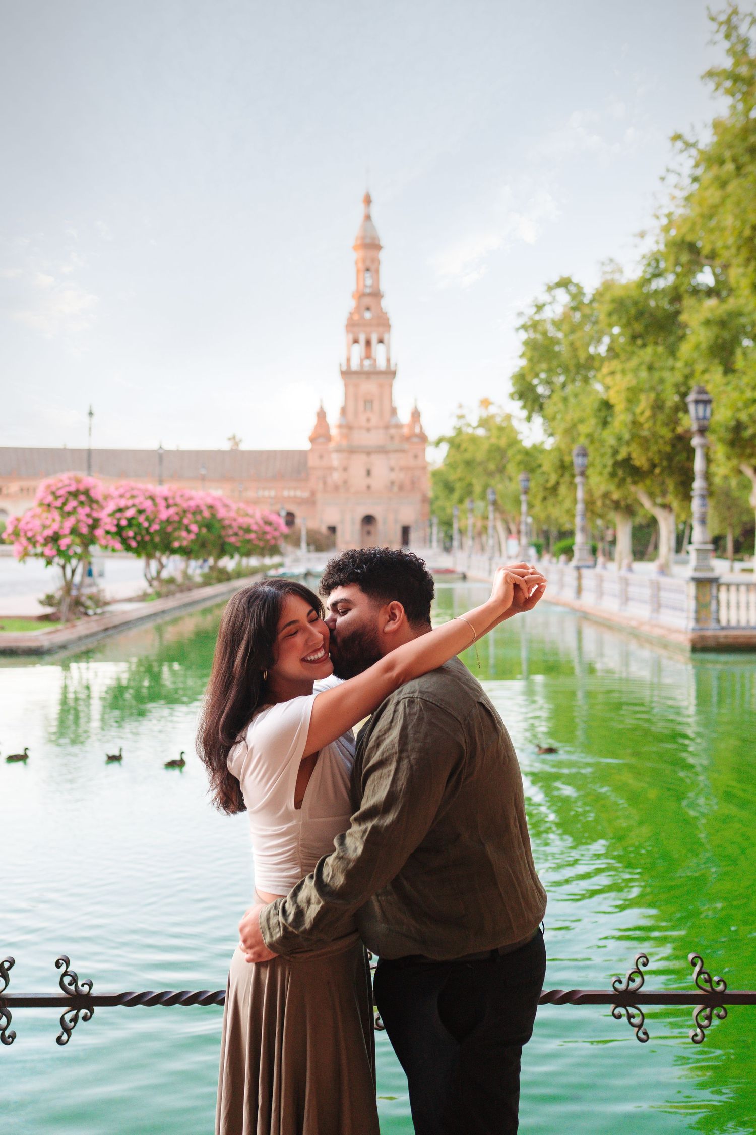 Couple embracing and laughing by a green canal at Plaza de España, Seville, with a historic tower backdrop.