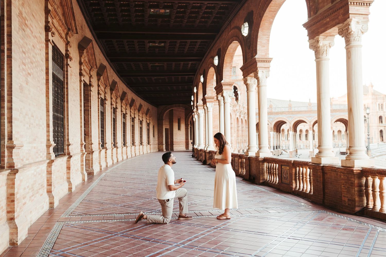 Man proposes on one knee to woman in white dress in Plaza de España arcade colonnade.