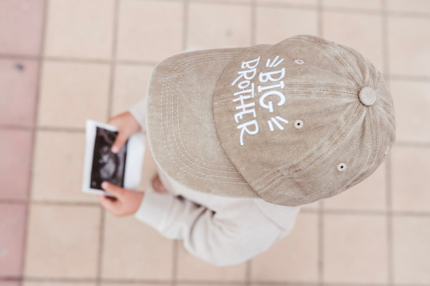 Child wearing Big Brother cap holding ultrasound photo from above.