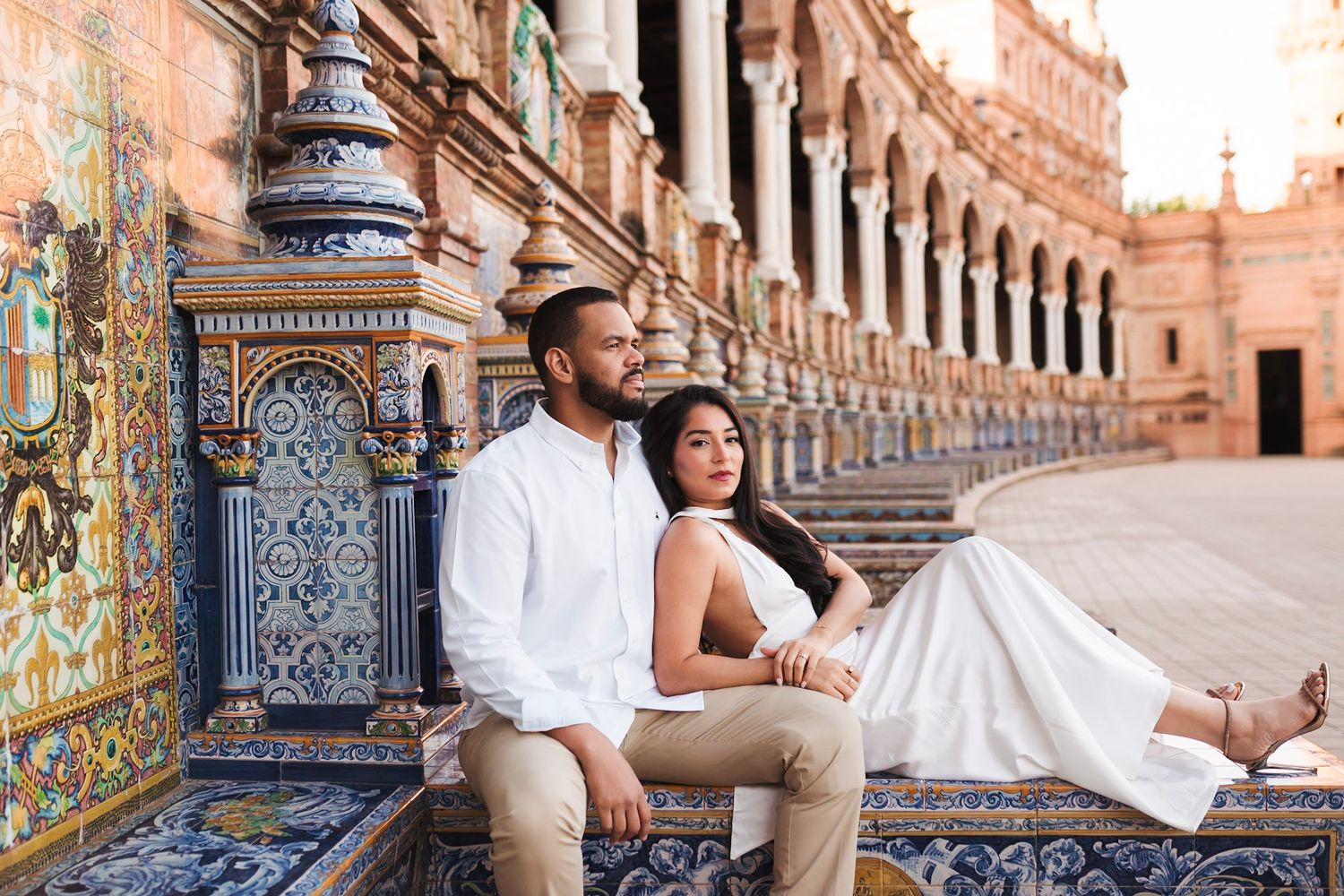 Couple sitting on ornate tiled bench at Plaza de Espana in Seville, woman in white dress leaning on man.