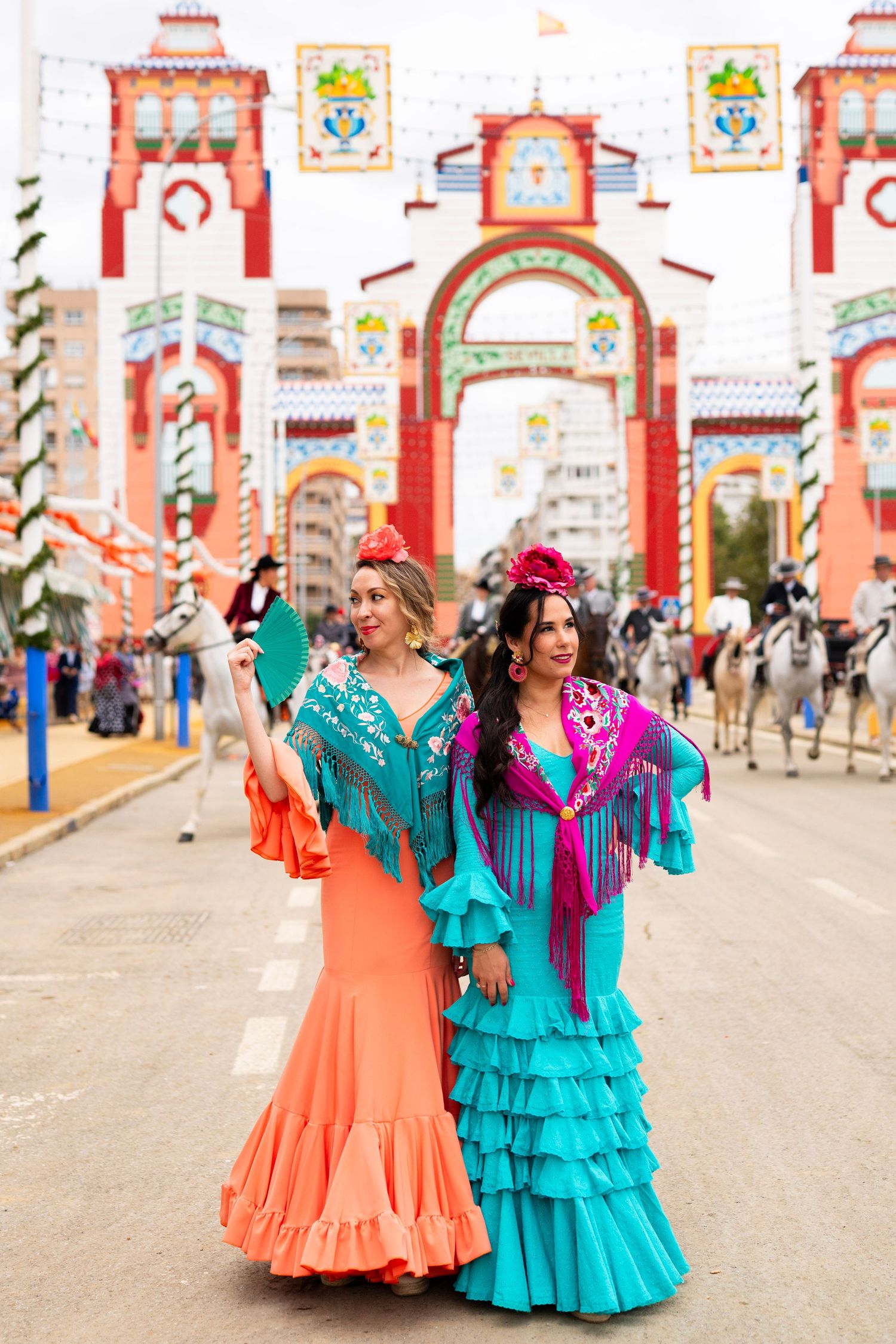 Two women in colorful flamenco dresses pose before ornate gate at Seville's Feria de Abril festival.