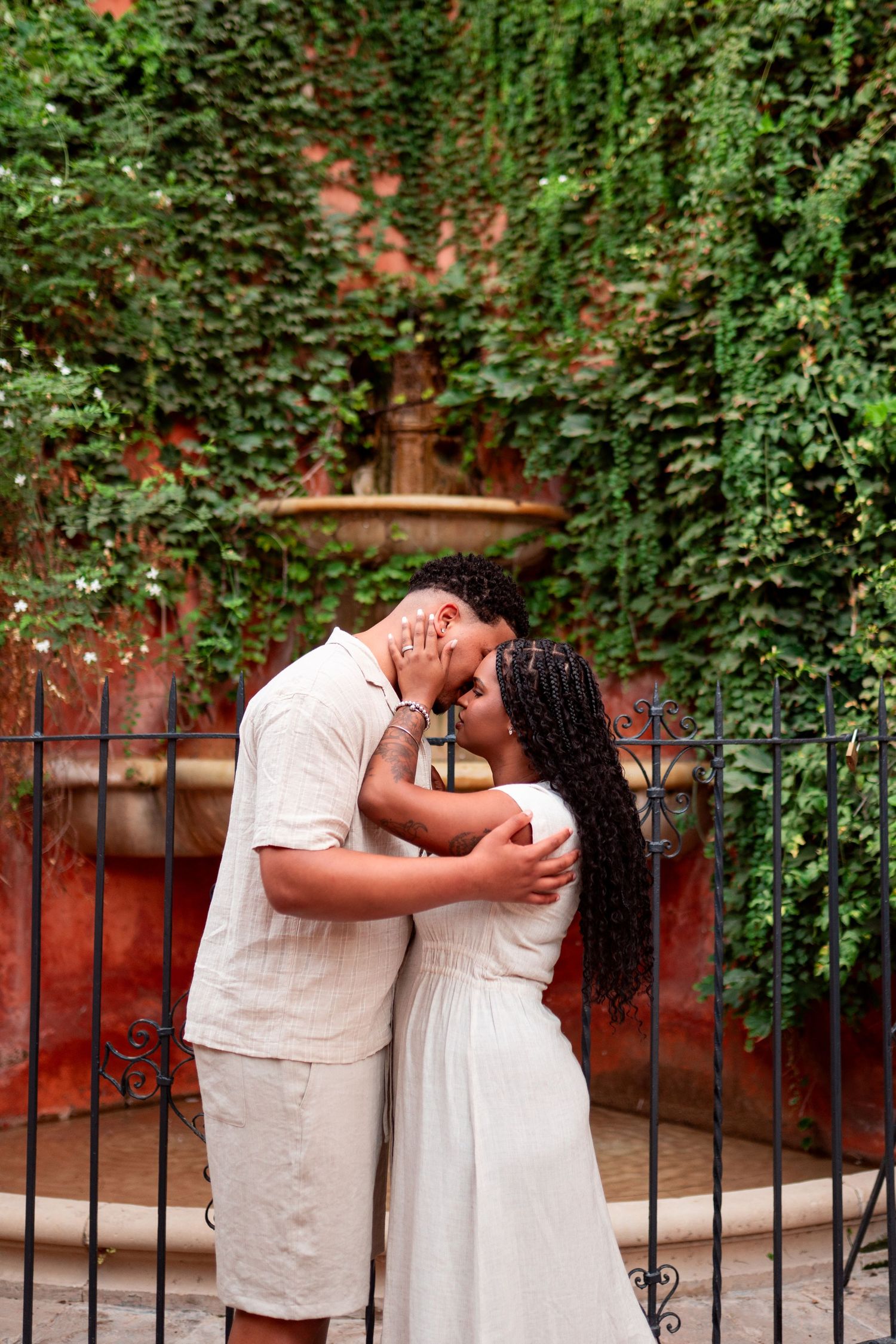 A couple shares a romantic kiss near a stone fountain surrounded by lush green ivy on a red wall.