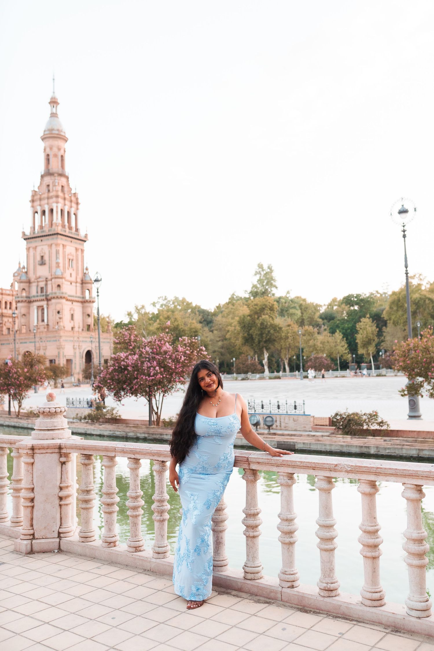 Pregnant woman in blue lace dress poses at Plaza de España, Seville, with ornate tower and canal backdrop.
