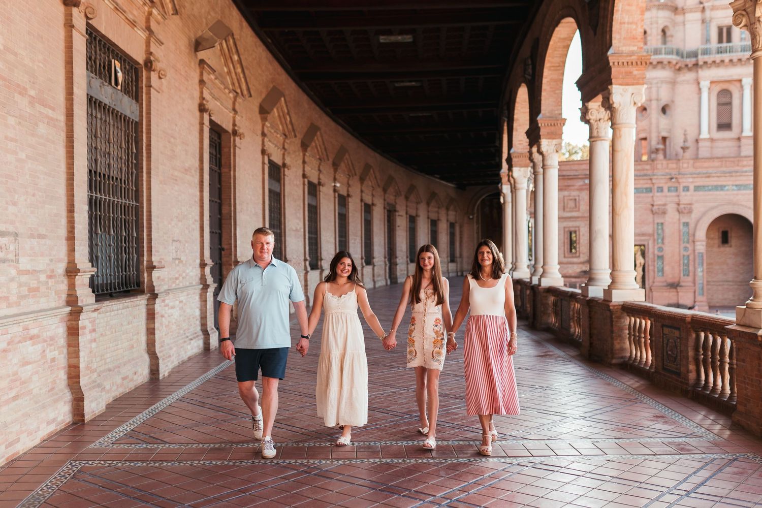 Family of four walking hand in hand through ornate Spanish plaza archway corridor.