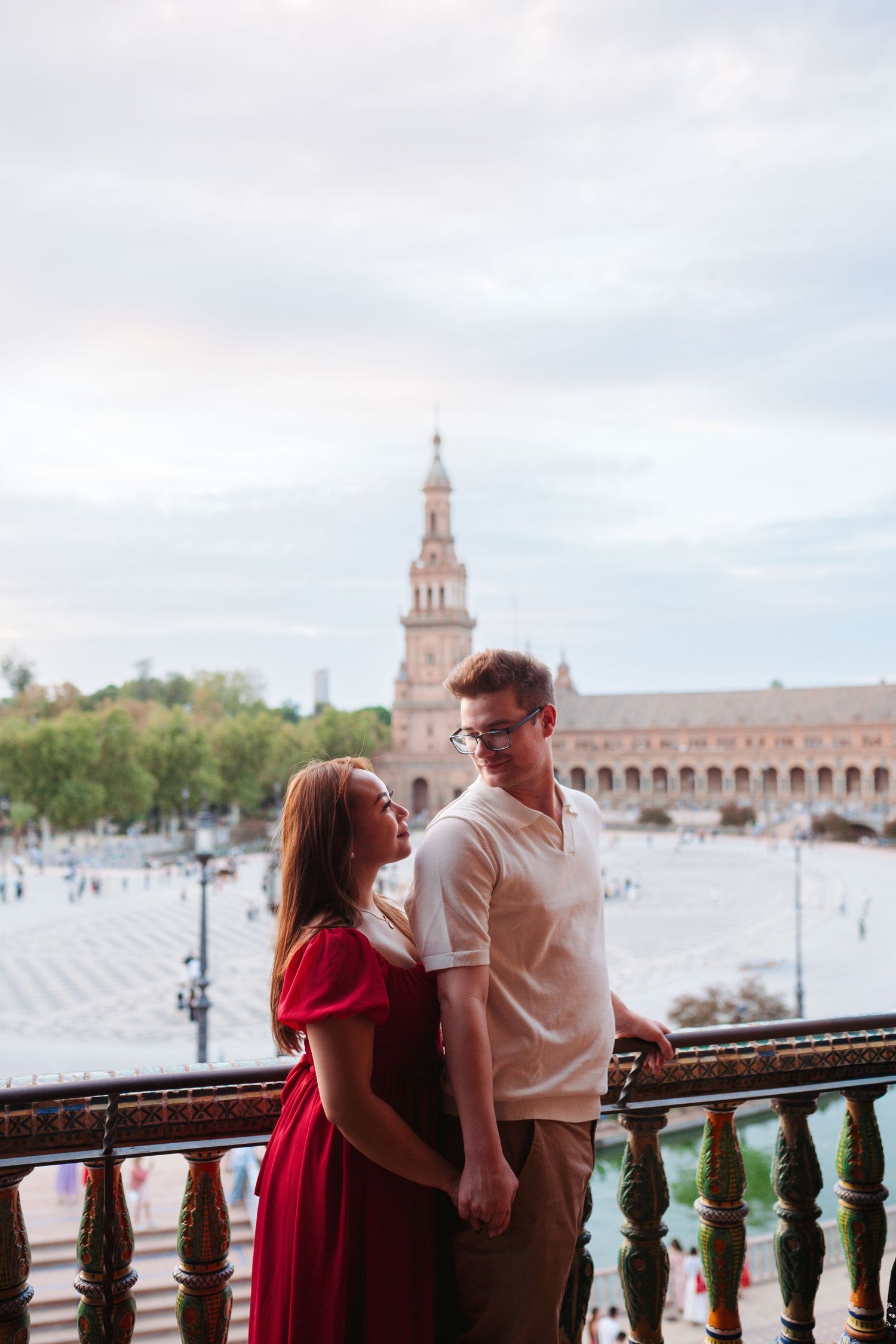 Couple holding hands on a balcony at Plaza de España in Seville, woman in red dress gazing at man in white shirt.