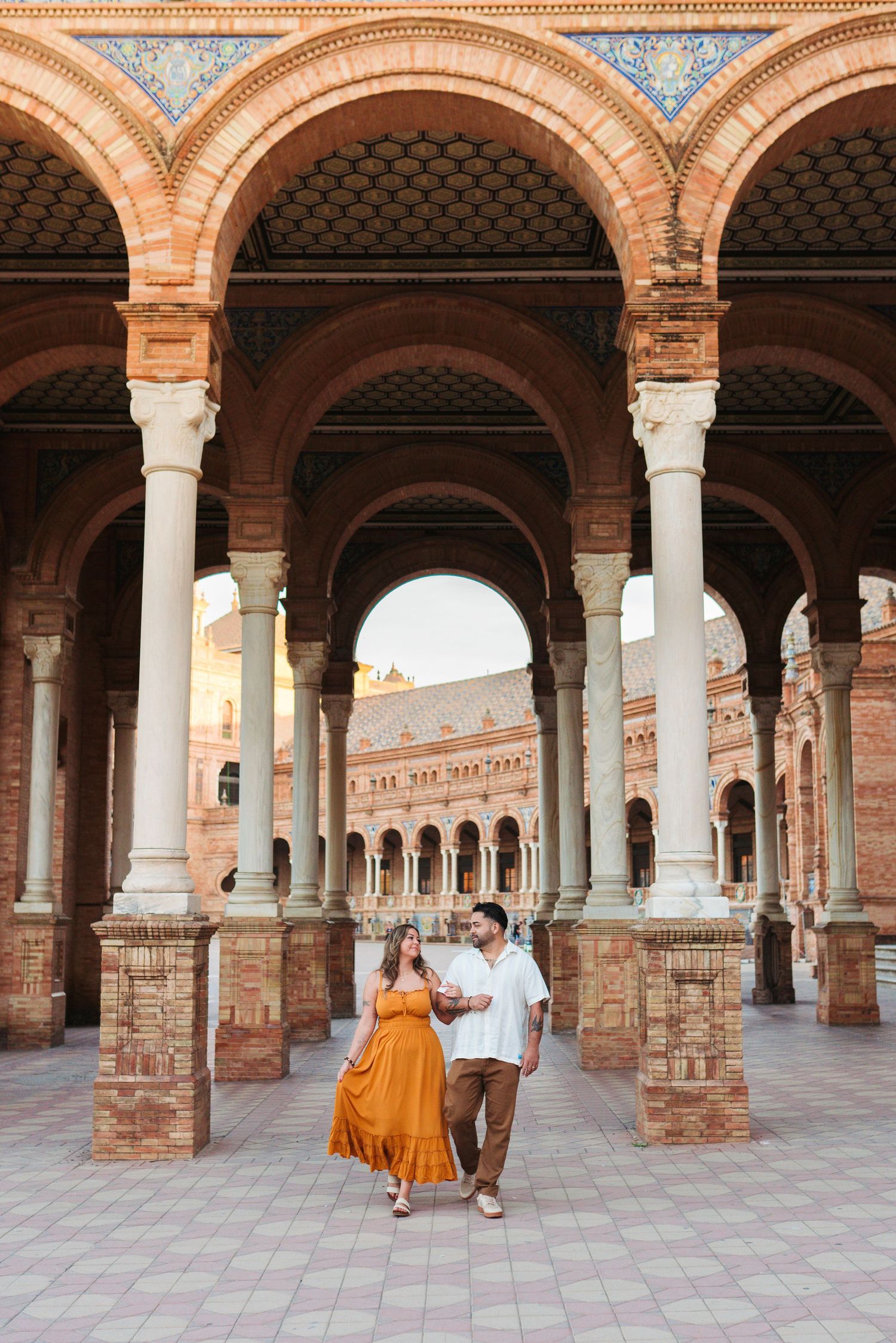 Couple walking under ornate brick arches at Plaza de España, Seville, woman in orange dress, man in white shirt.