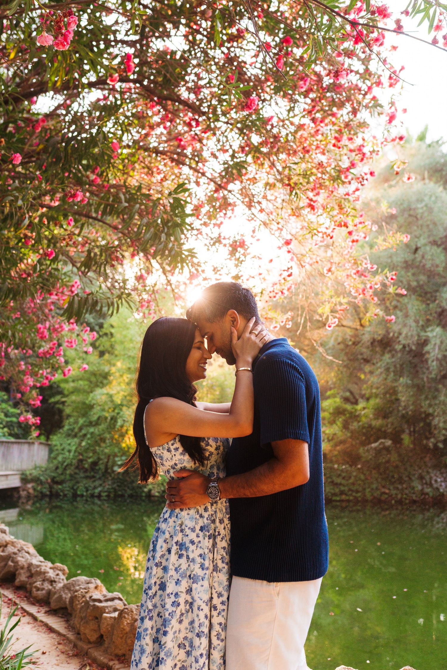 Romantic couple sharing a tender moment under blooming pink flowers by a serene garden pond in warm sunlight.