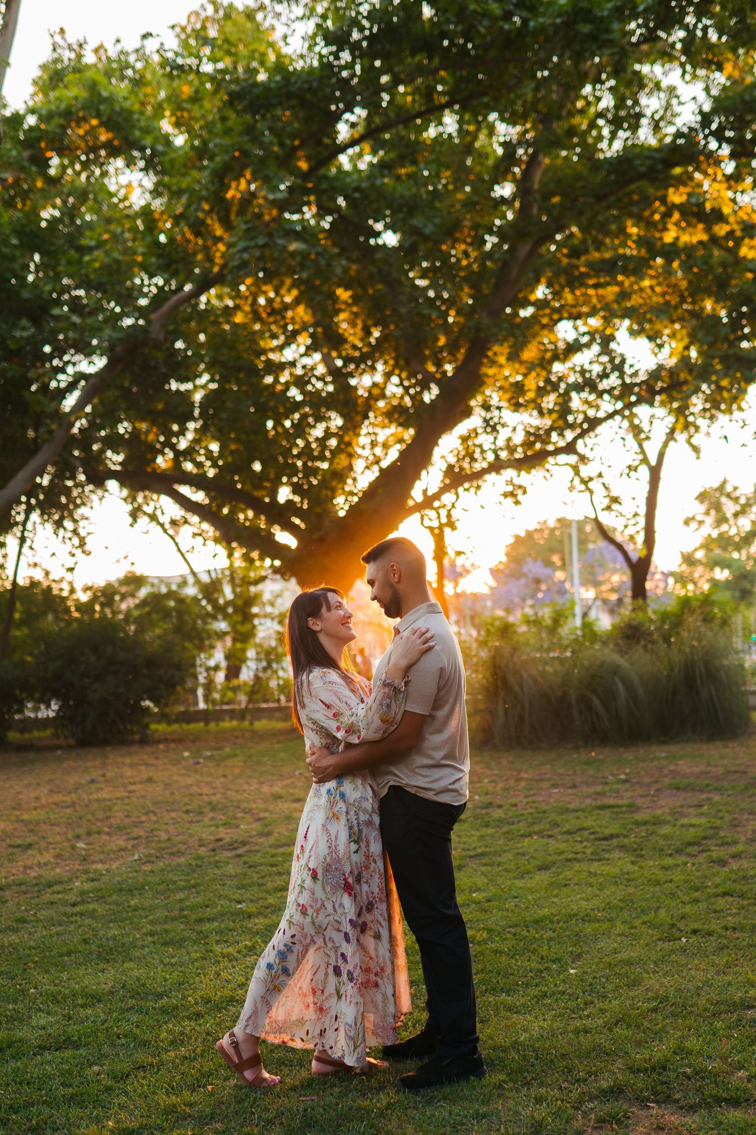 Couple embracing at golden hour sunset in a lush green park, woman in floral dress, romantic outdoor portrait.