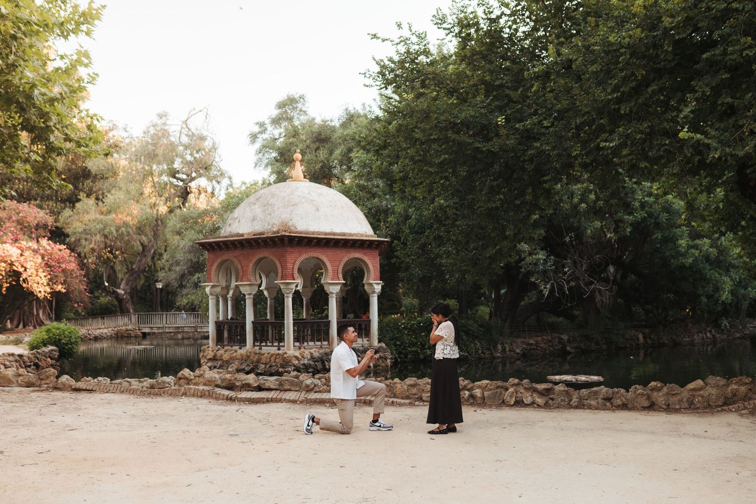 Man proposing to woman on one knee in a scenic park with ornate pavilion and lush trees in background.