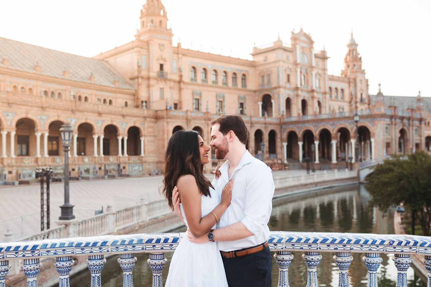 Couple embracing on tiled bridge at golden hour with Plaza de España grand facade reflected in canal.