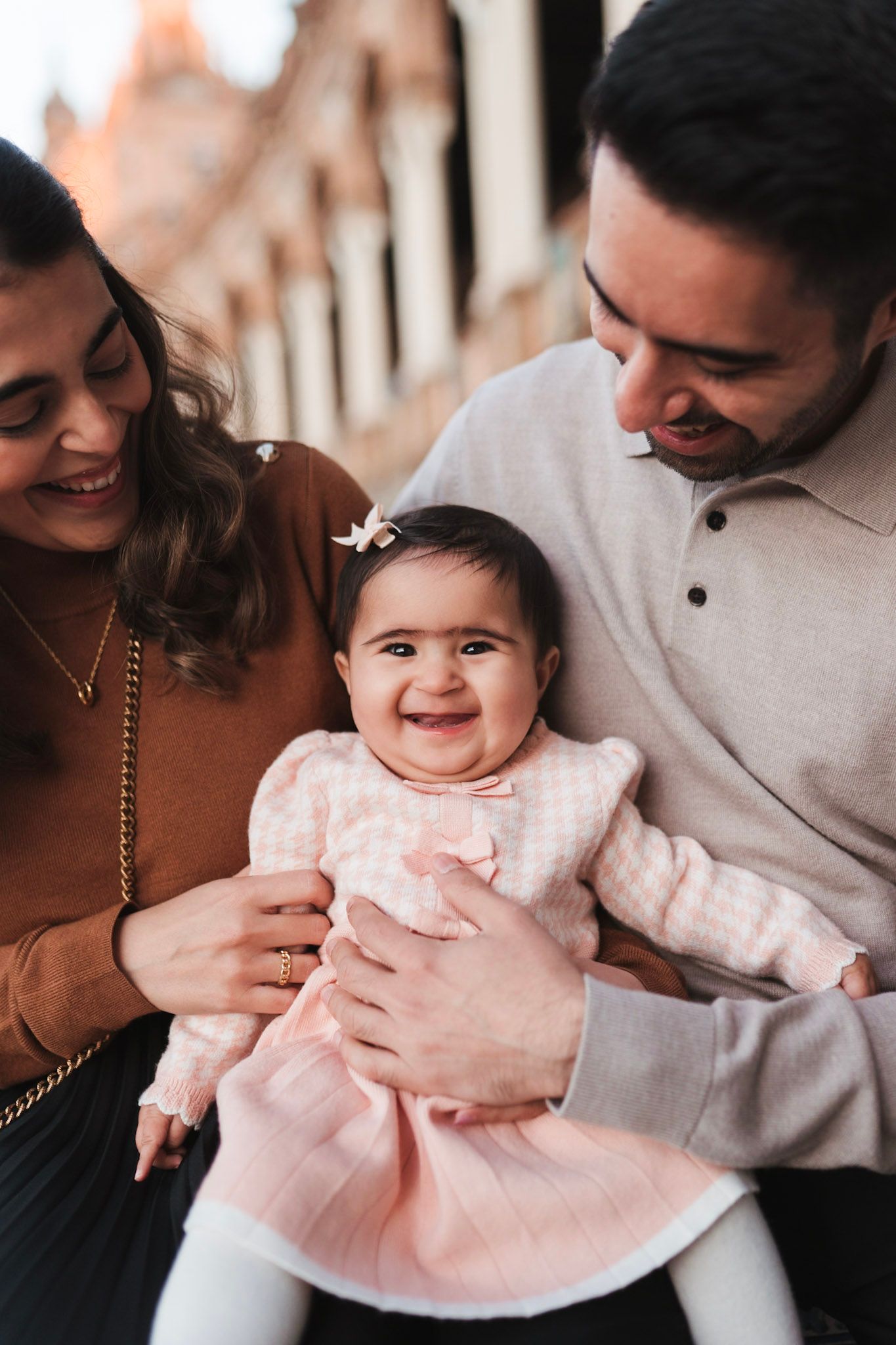 Proud parents smiling at their laughing baby girl in a pink dress at Plaza de España, Seville.