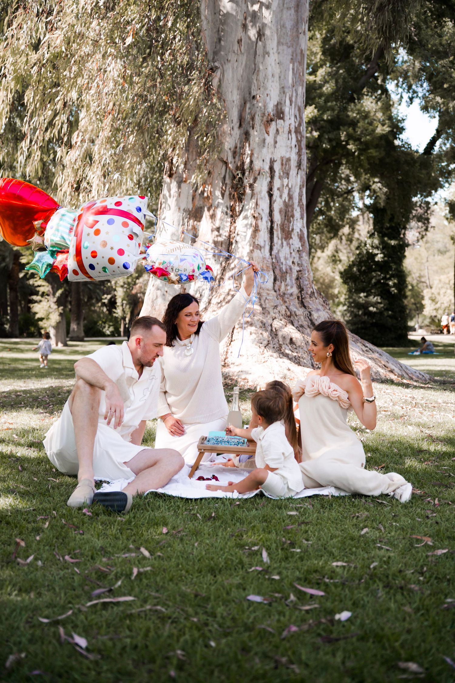 Joyful family in white outfits celebrates with colorful balloons during a picnic under a large tree.