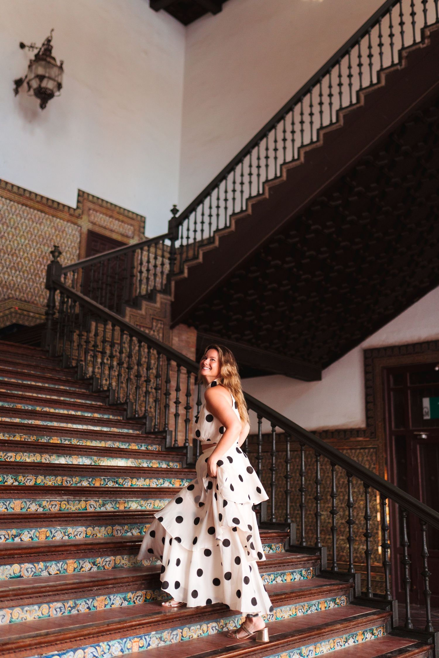 Woman in white polka dot dress posing on ornate tiled staircase with dark wood railings in elegant interior.