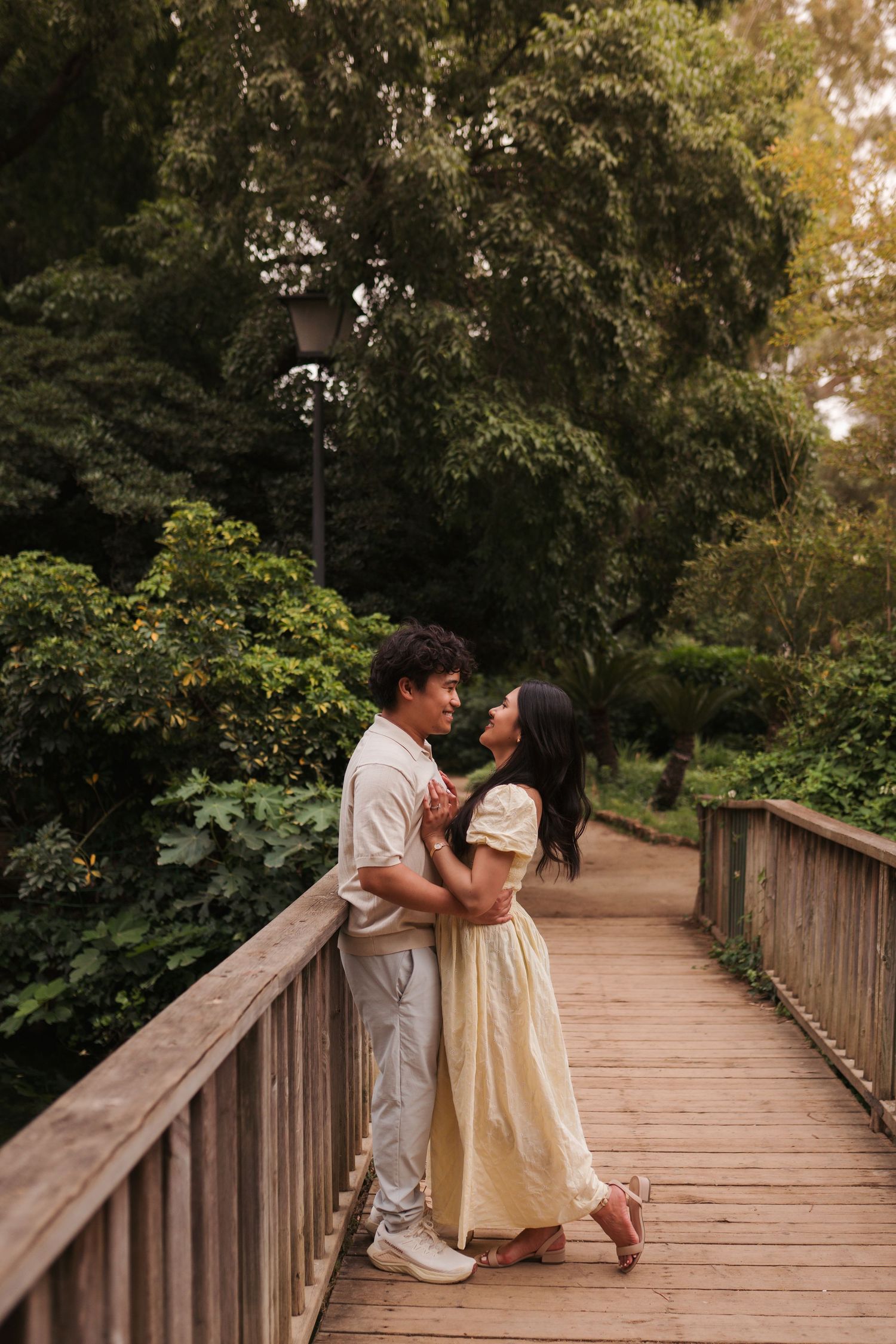 Couple embracing on a wooden bridge surrounded by lush green trees in a romantic park setting.