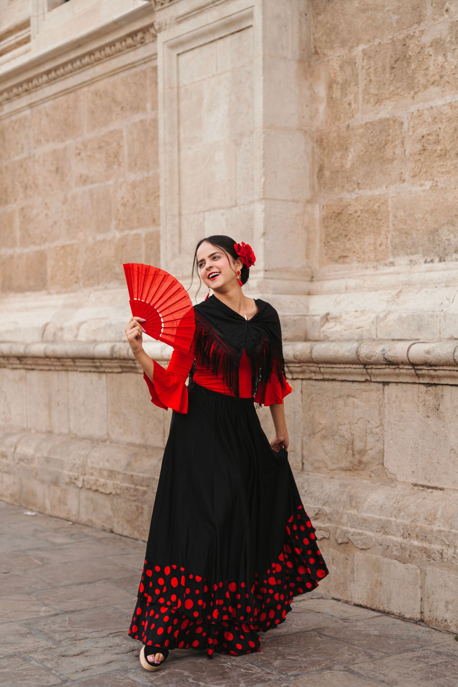 Flamenco dancer in black polka dot dress holds red fan against stone building.