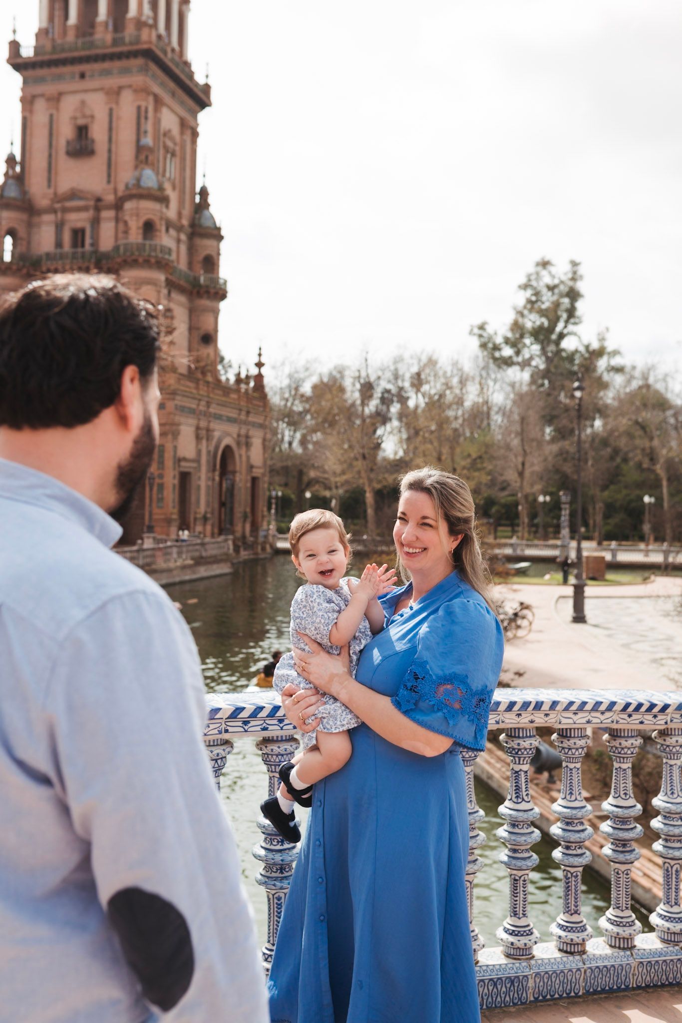 A woman in a blue dress holds her baby while smiling at her partner by the canal at Plaza de España.