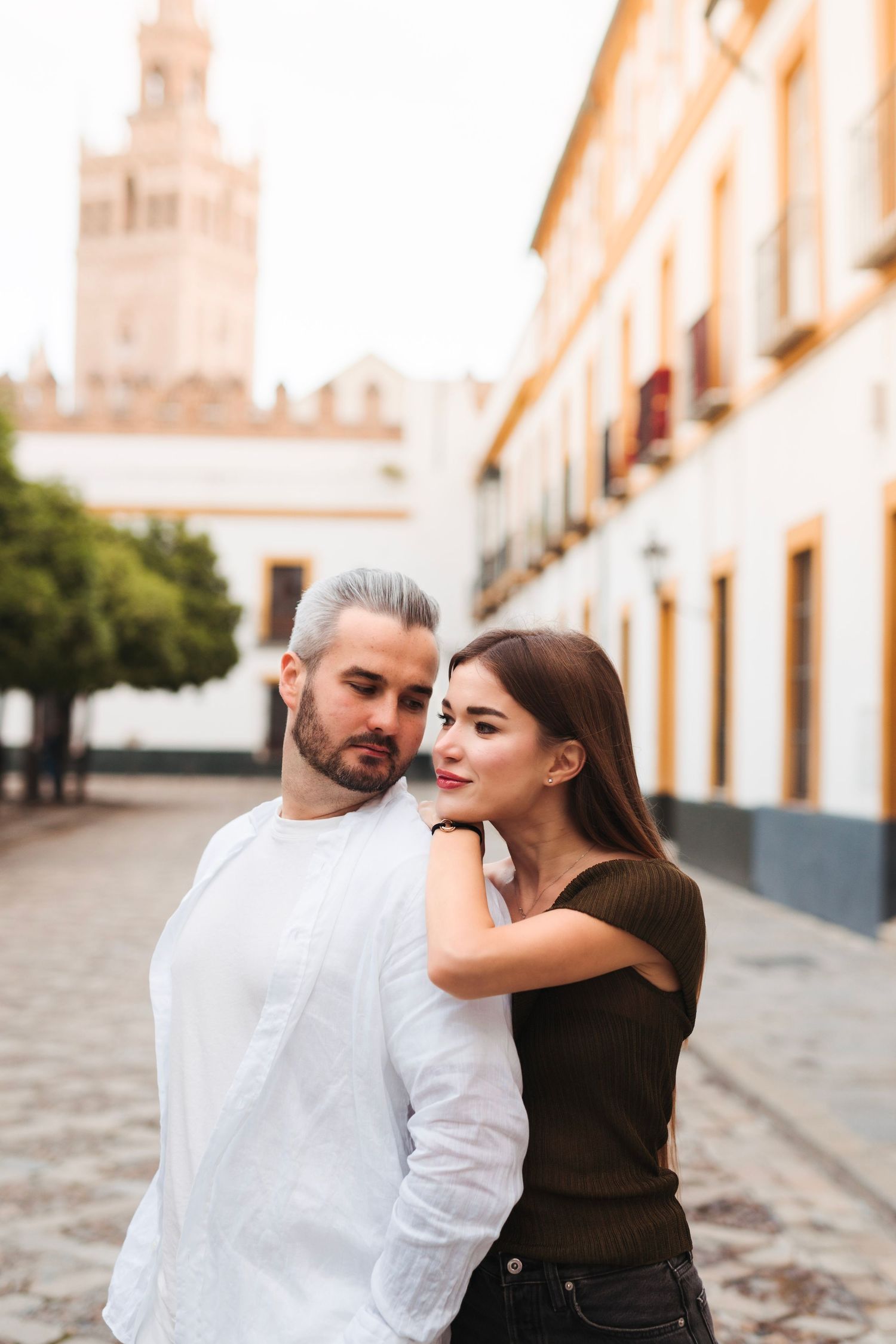 Couple embracing on cobblestone street with Giralda tower visible in background in Seville, Spain.