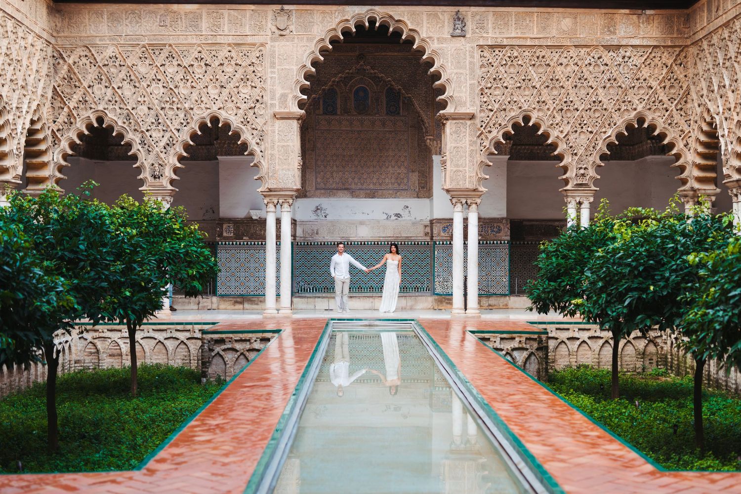 Couple holding hands by a reflective pool in the ornate Moorish courtyard of Real Alcazar, Seville.