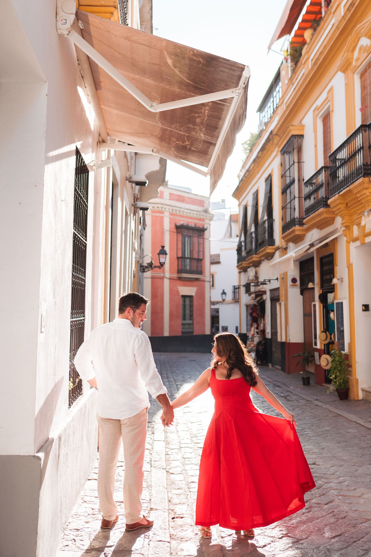 Couple holding hands walking through a charming cobblestone street, woman in flowing red dress, man in white outfit.