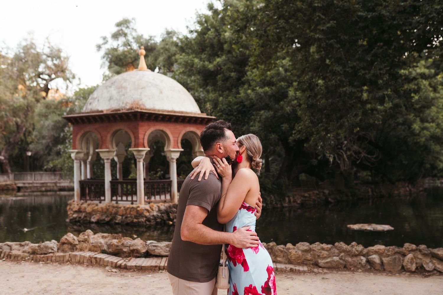 Couple embracing and kissing near an ornate gazebo by a pond in a lush garden setting.
