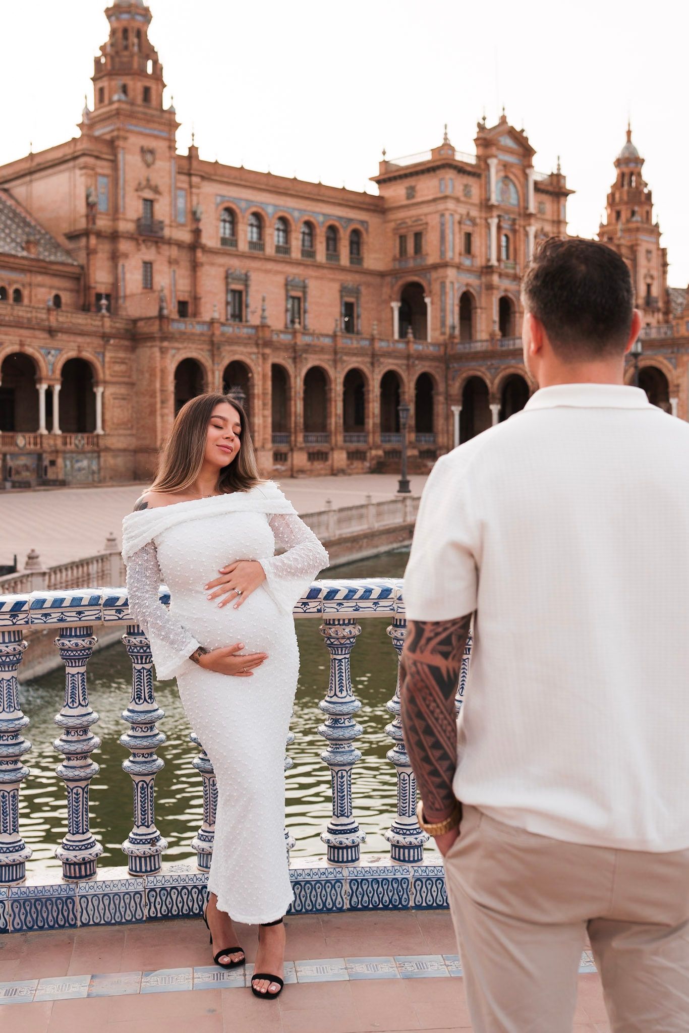 Couple at Plaza de España Seville, pregnant woman in white lace dress faces tattooed partner by canal.