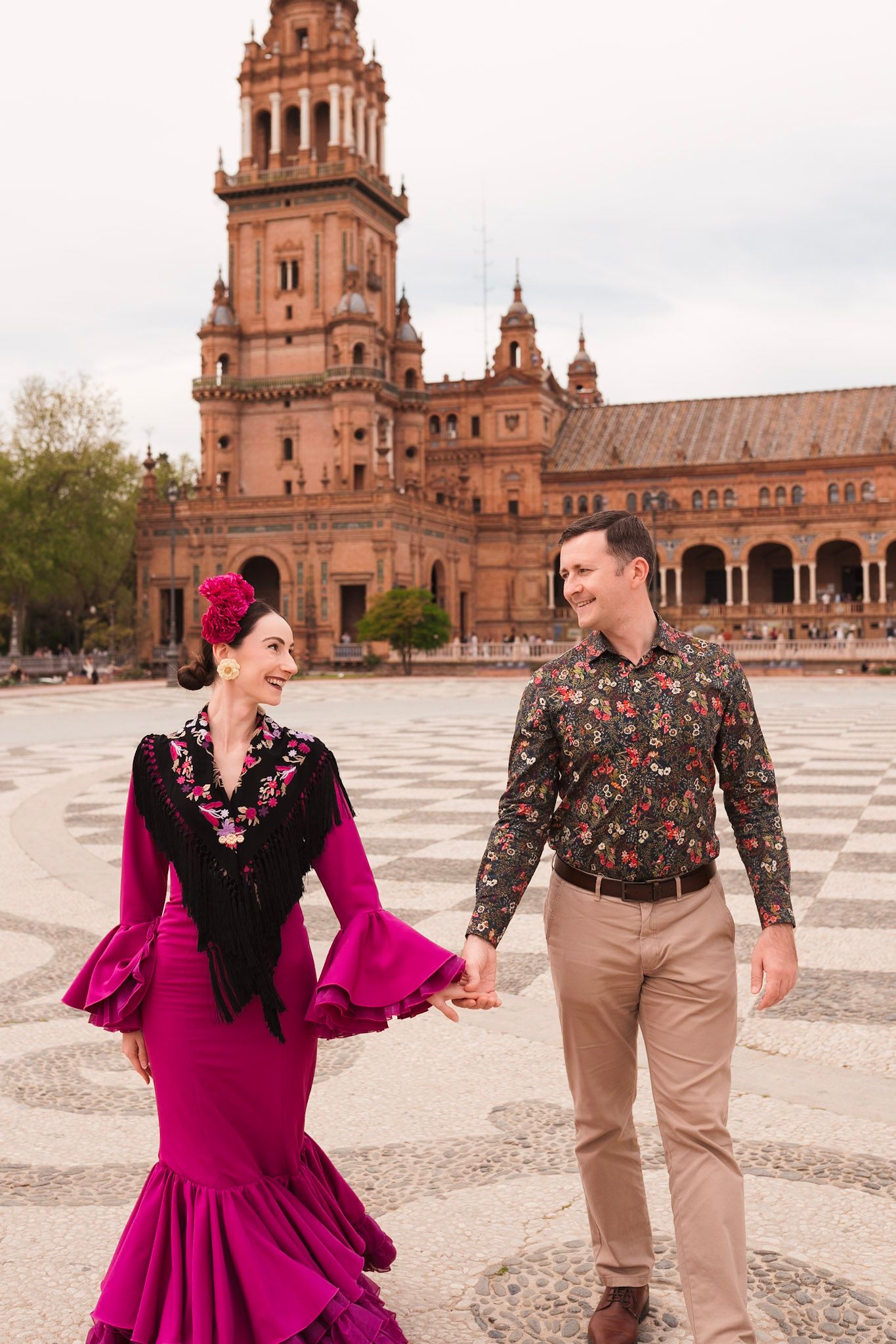 Couple holding hands at Plaza de España Seville, woman in magenta flamenco dress with black shawl, man in floral shirt.