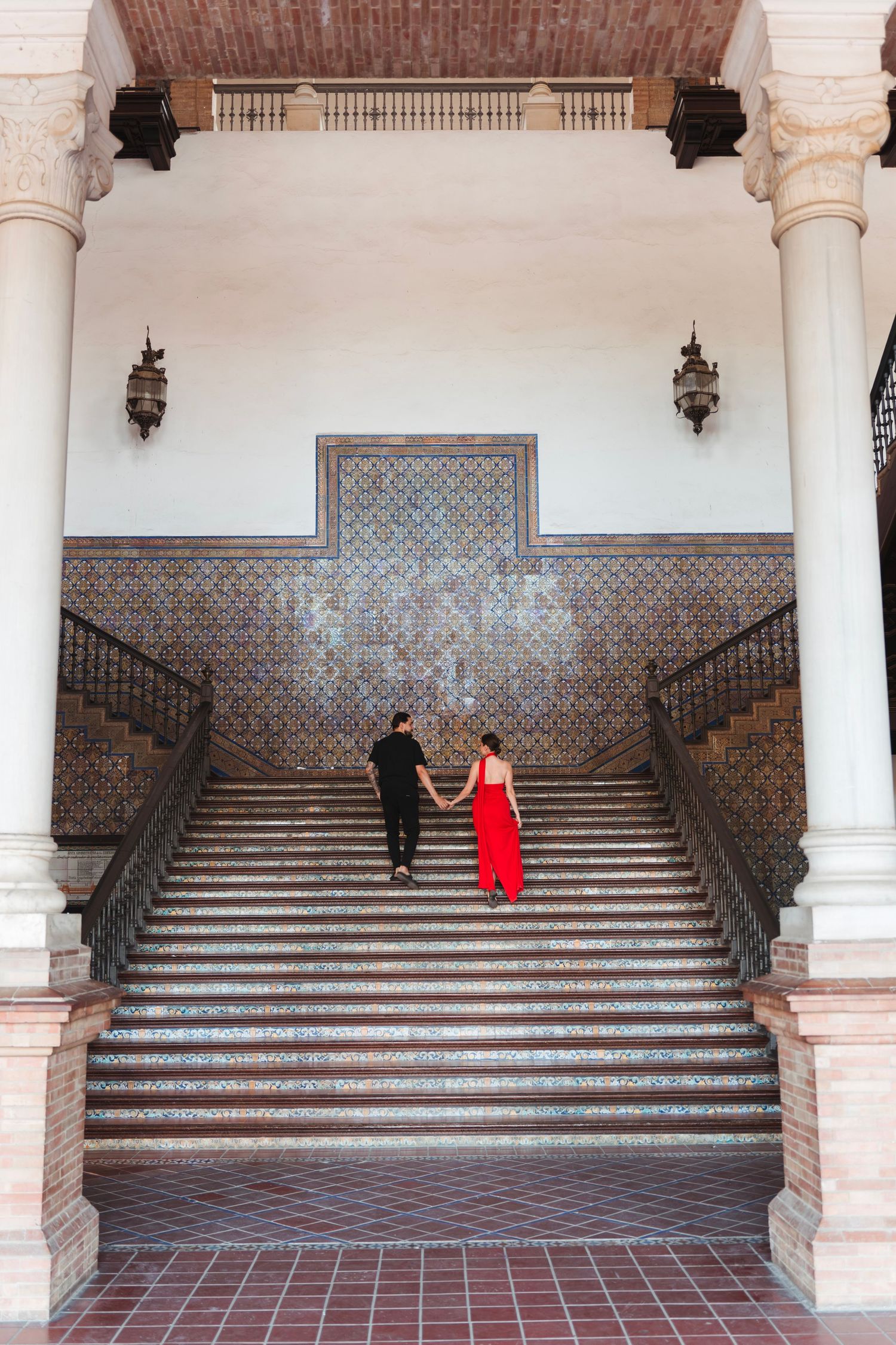 Couple holding hands ascending grand mosaic staircase with ornate tile walls in Seville historic building.