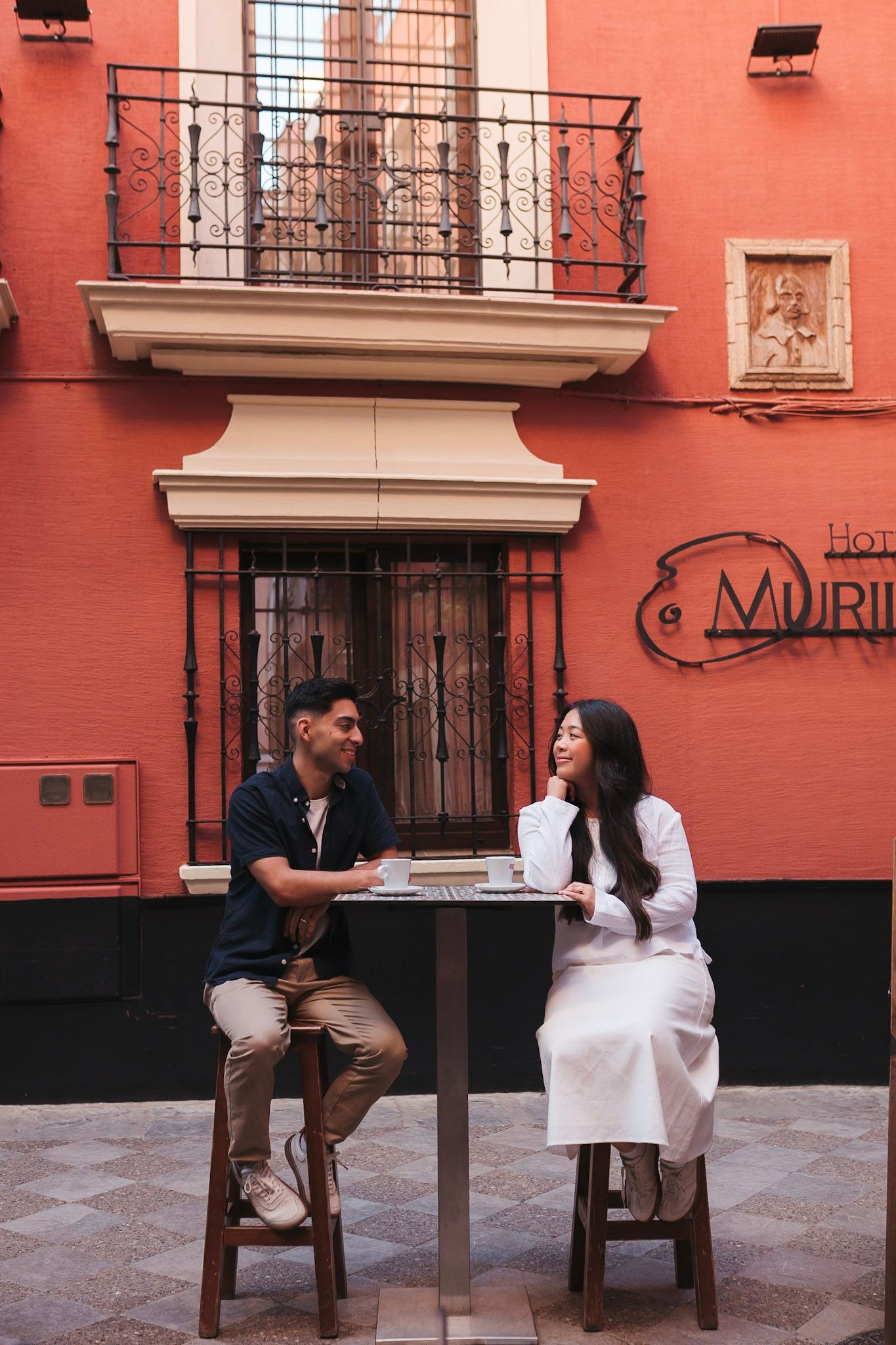 Couple enjoying coffee at an outdoor cafe beside a red building in Seville, Spain.