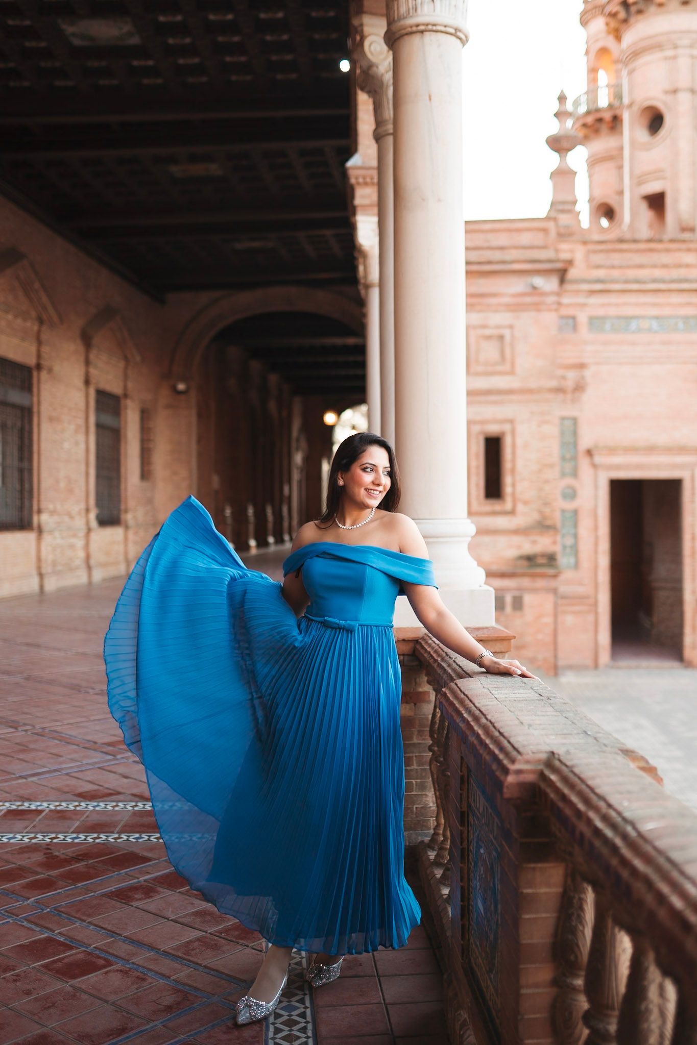 Woman in flowing blue off-shoulder gown poses at Plaza de España colonnade.