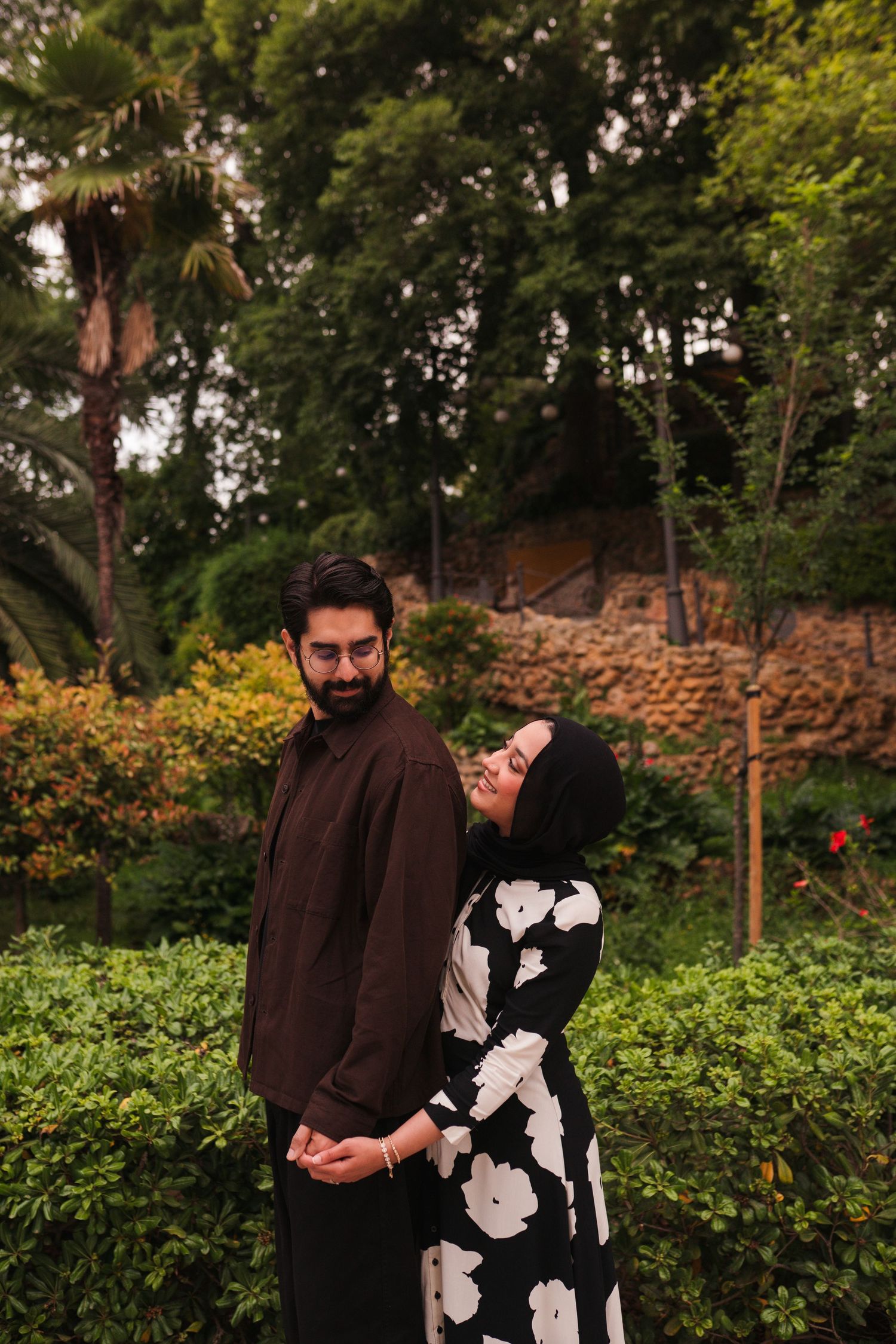 Couple in garden, man in brown kurta, woman in floral hijab dress, holding hands among lush greenery.