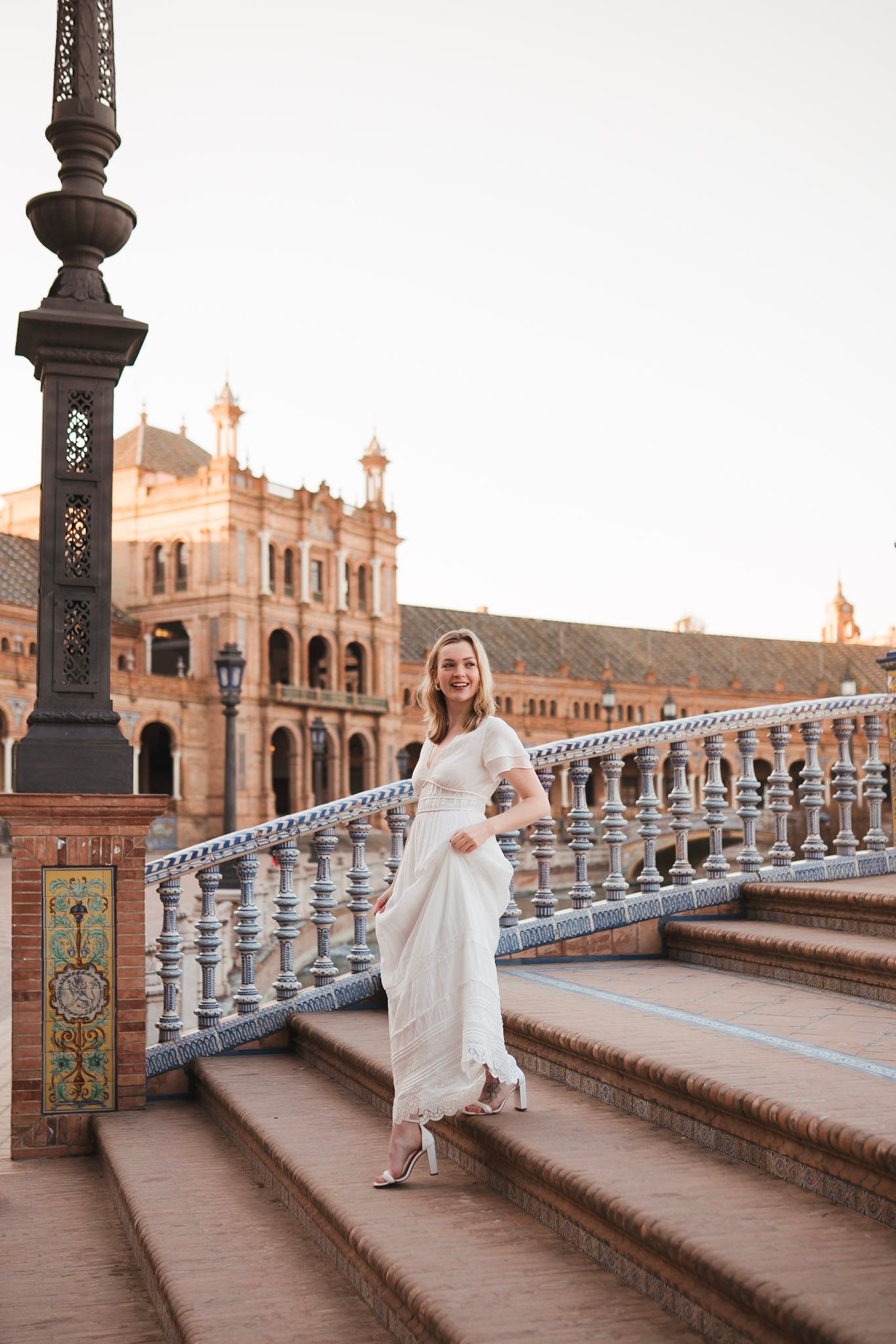 Blonde woman in white gown walks Plaza de España bridge at sunset.