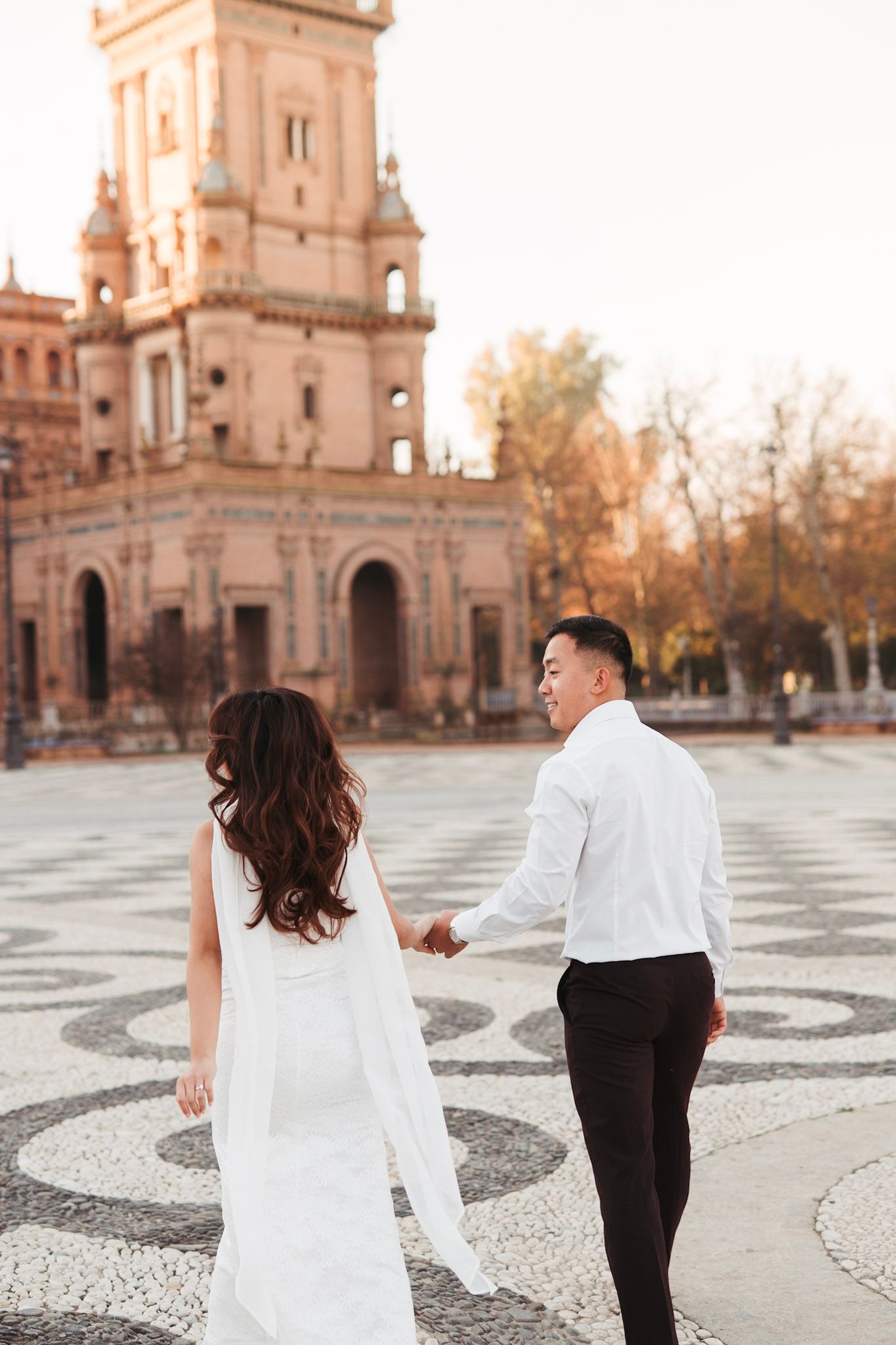 Bride and groom holding hands walking toward ornate Spanish plaza tower at golden hour sunset.