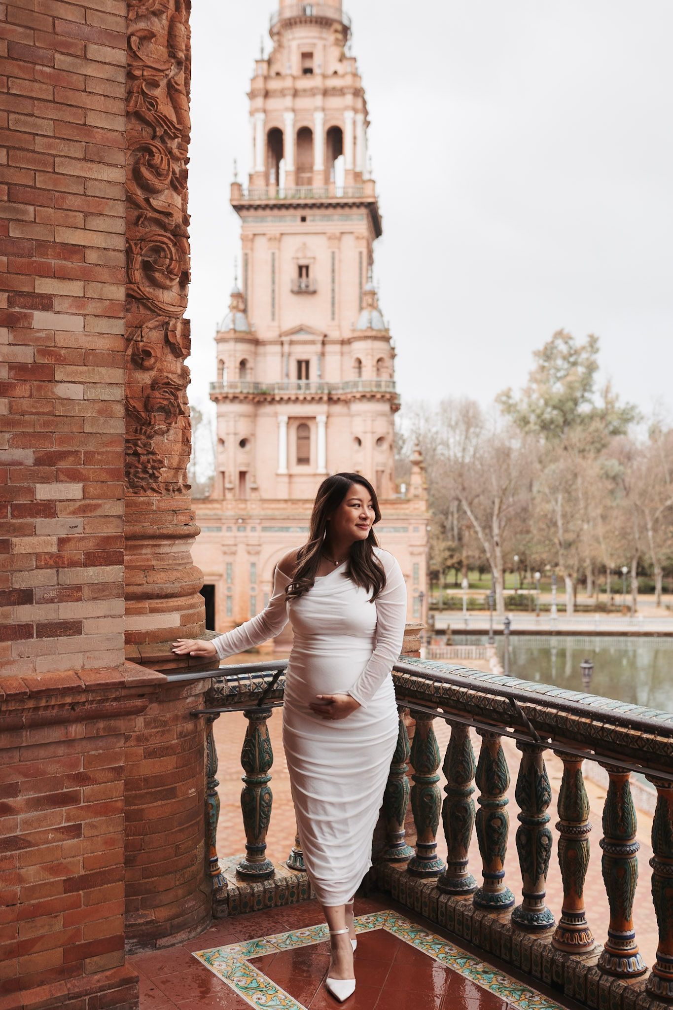 Pregnant woman in white dress poses on balcony at Plaza de España, Seville, with tower backdrop.