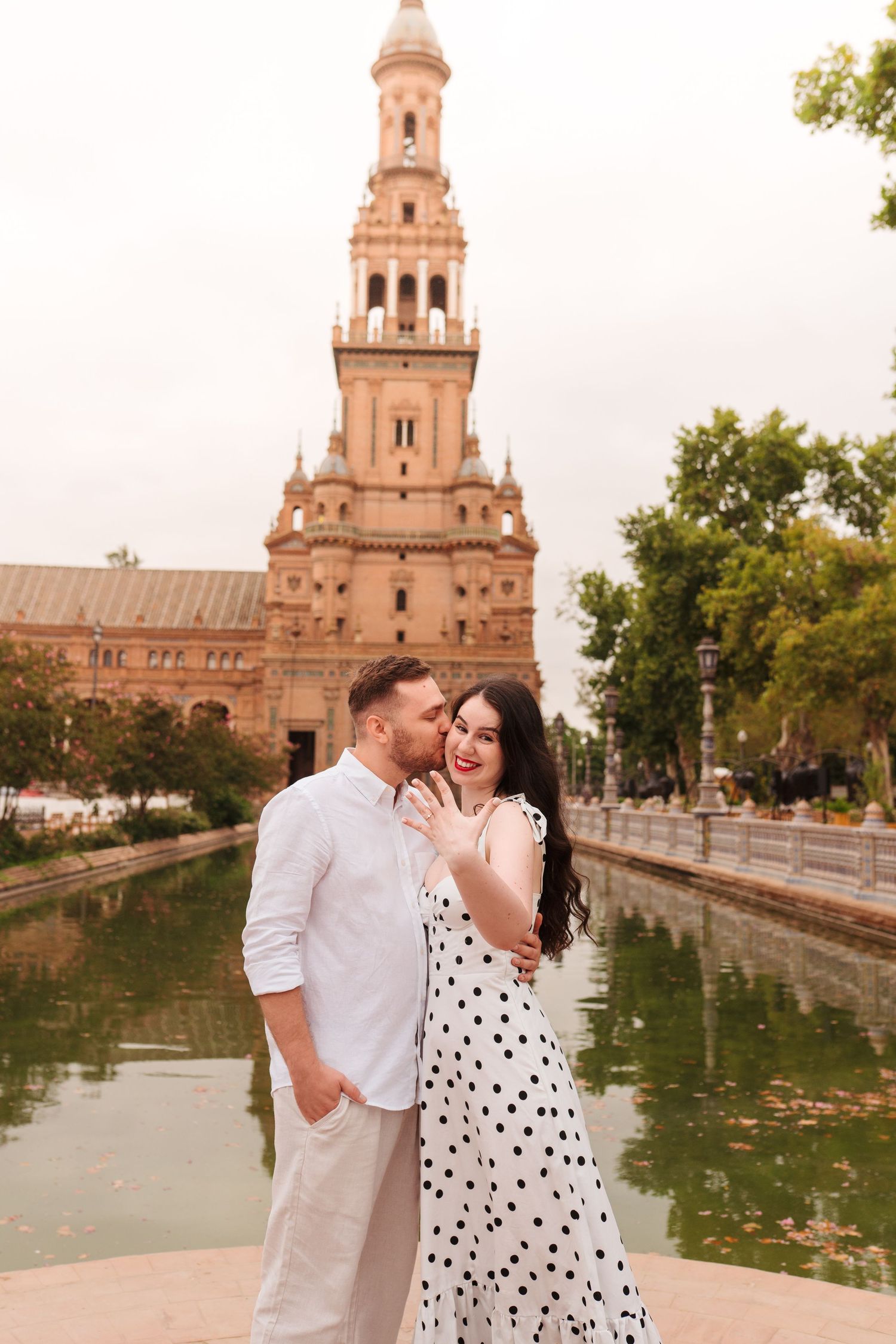 Couple embracing and kissing by a canal at Plaza de España, Seville, woman in polka dot dress.