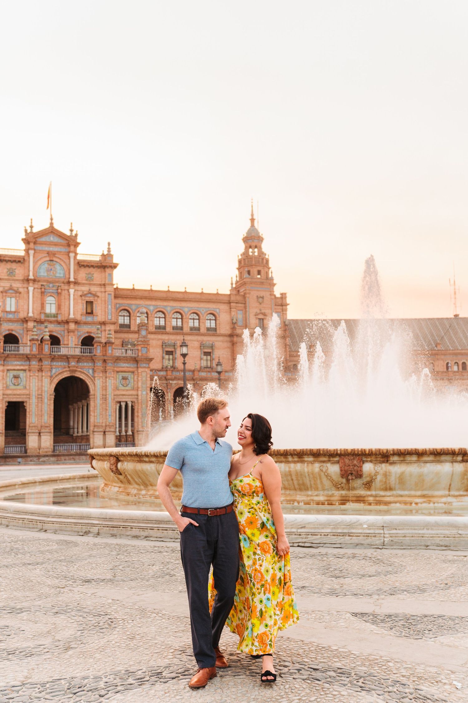 Couple embracing and smiling at Plaza de España in Seville, Spain, with fountain and ornate building at sunset.