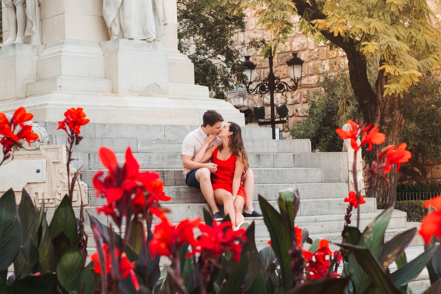 Romantic couple kissing on steps surrounded by vibrant red flowers near white statue.