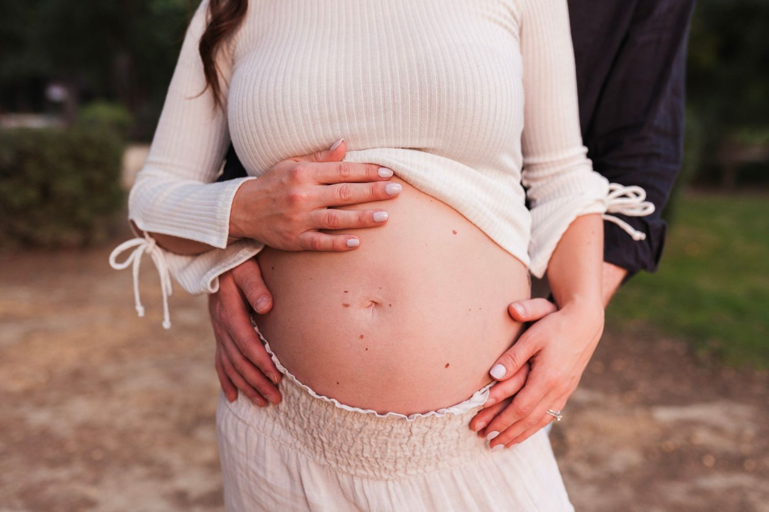 Close-up of pregnant woman's bare belly being cradled by couple's hands outdoors.