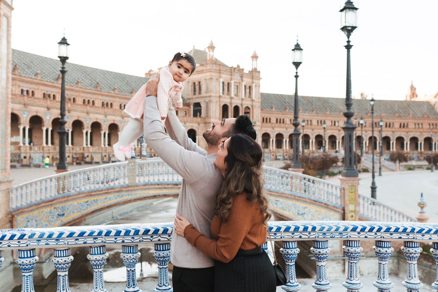 A man lifts a baby joyfully in the air while a woman watches at Plaza de España, Seville.