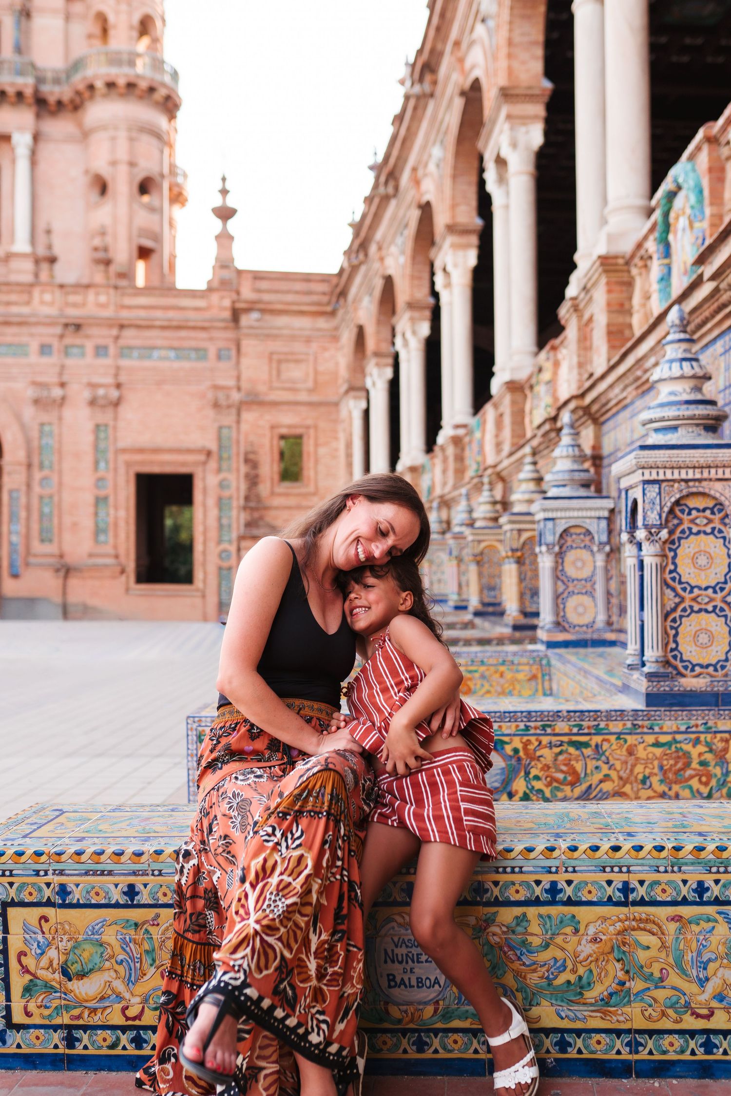 A mother and daughter share a joyful embrace on ornate tiled benches at Plaza de España, Seville, Spain.