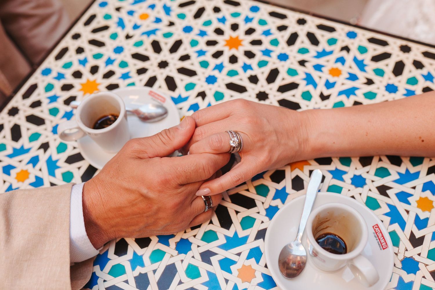 Couple holding hands with diamond ring on mosaic tile table with espresso cups.