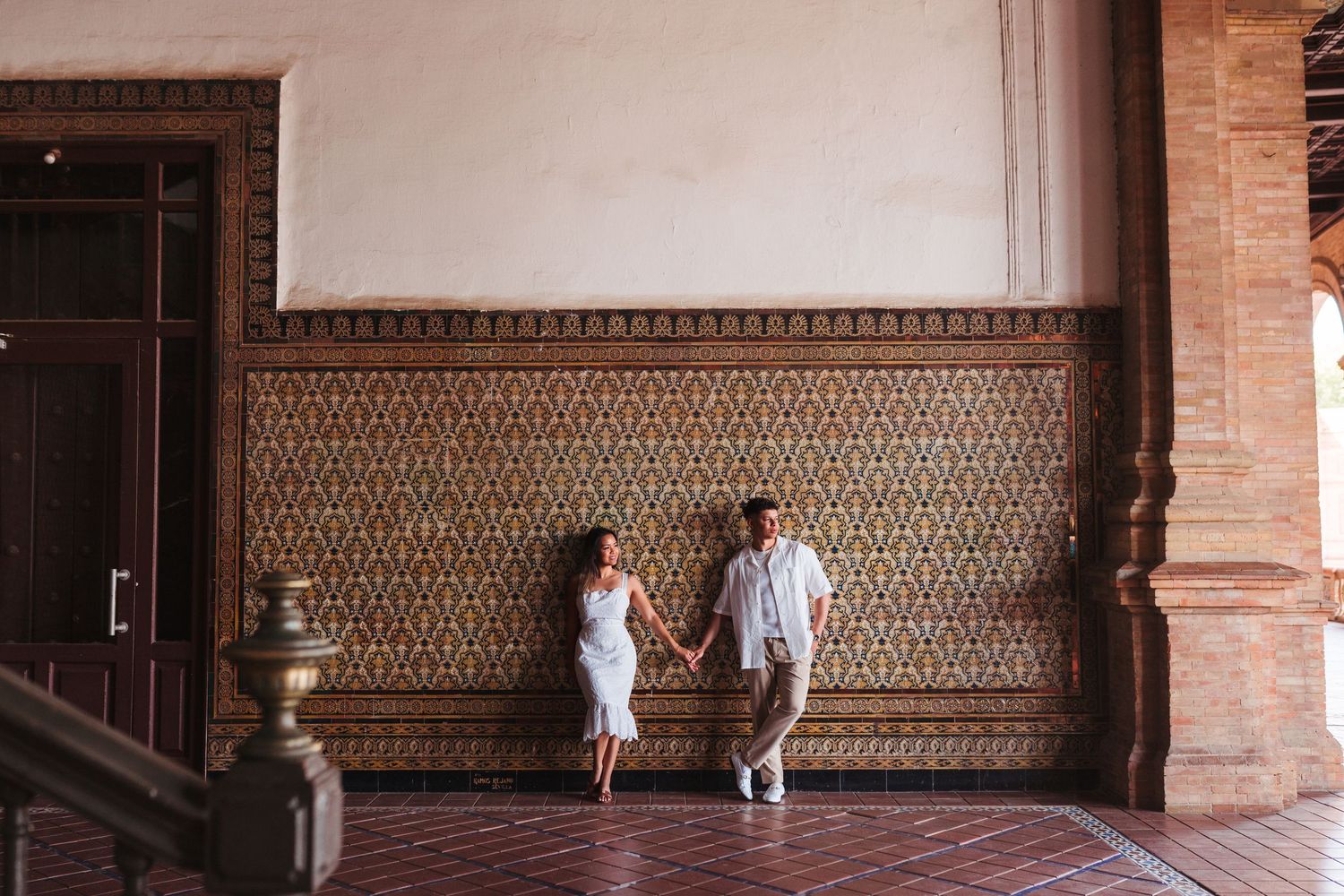 Couple walking hand in hand along ornate tiled wall inside Plaza de España Seville.