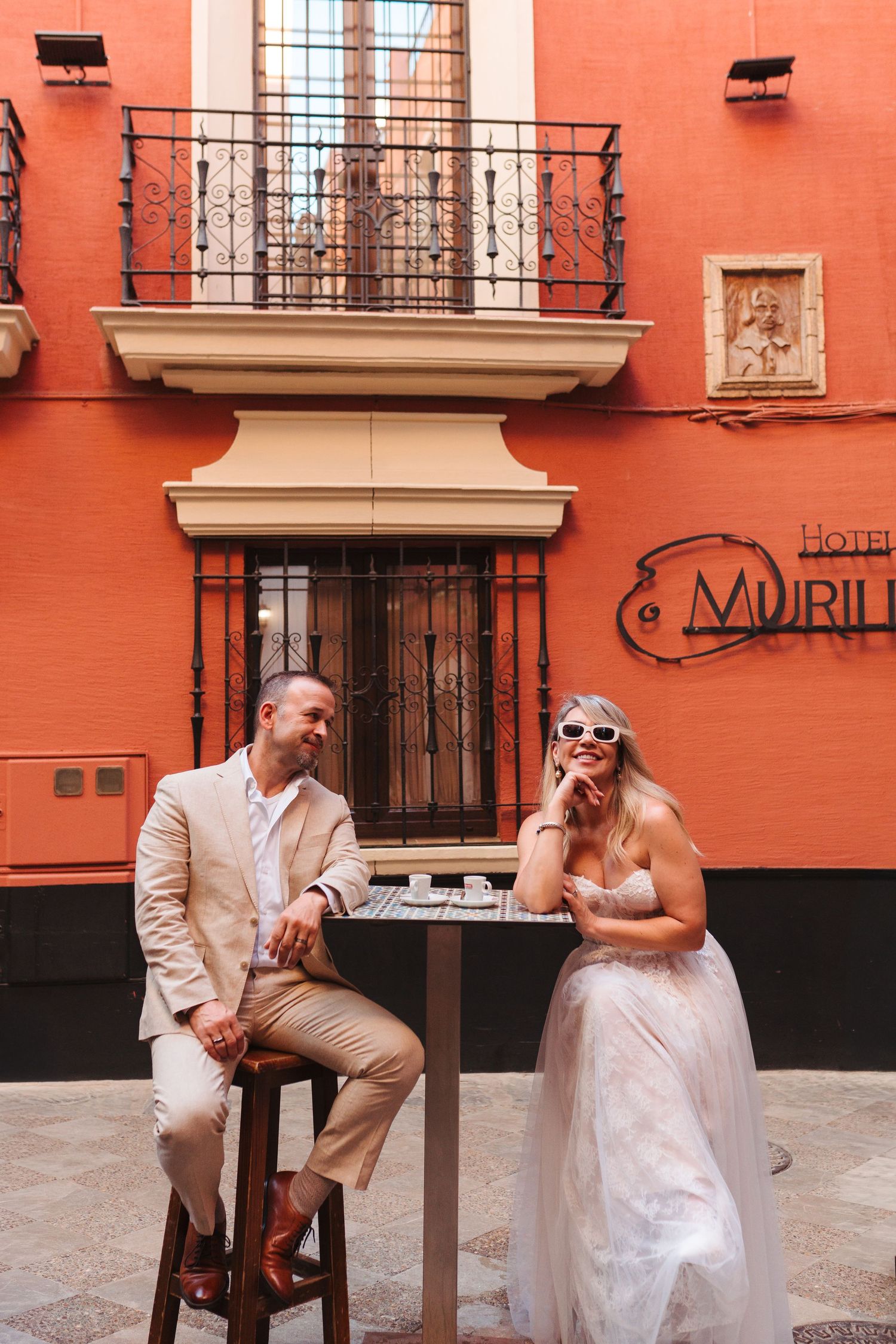 Bride and groom laughing at outdoor café table beside Hotel Murillo in Seville.